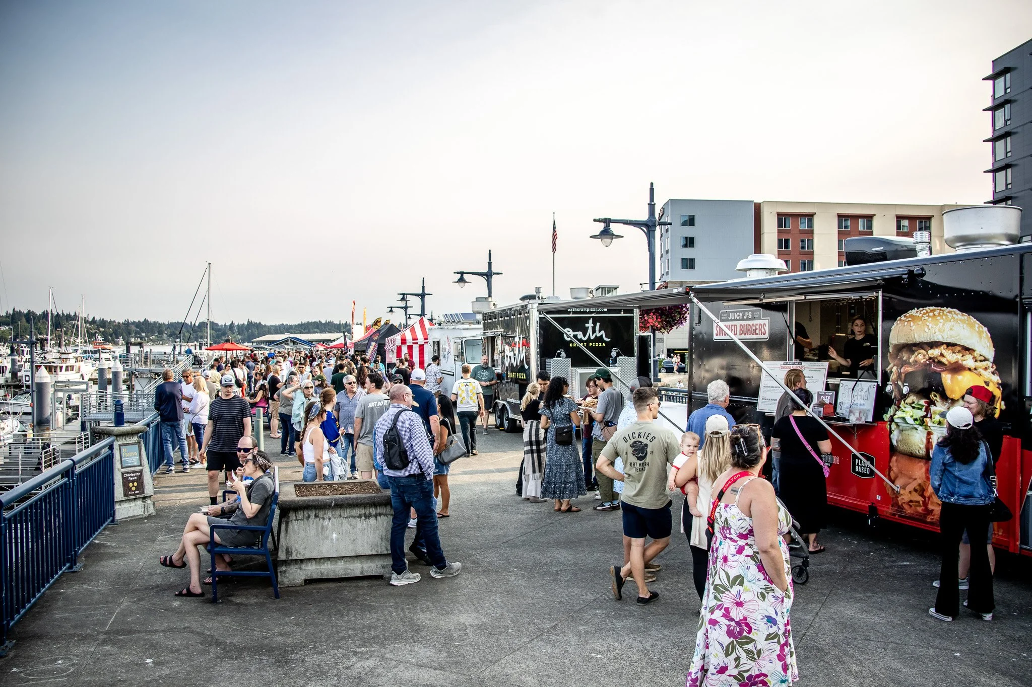 Crowd of people at a waterfront food festival with food trucks, boats in the background, and waterfront buildings under a clear sky.