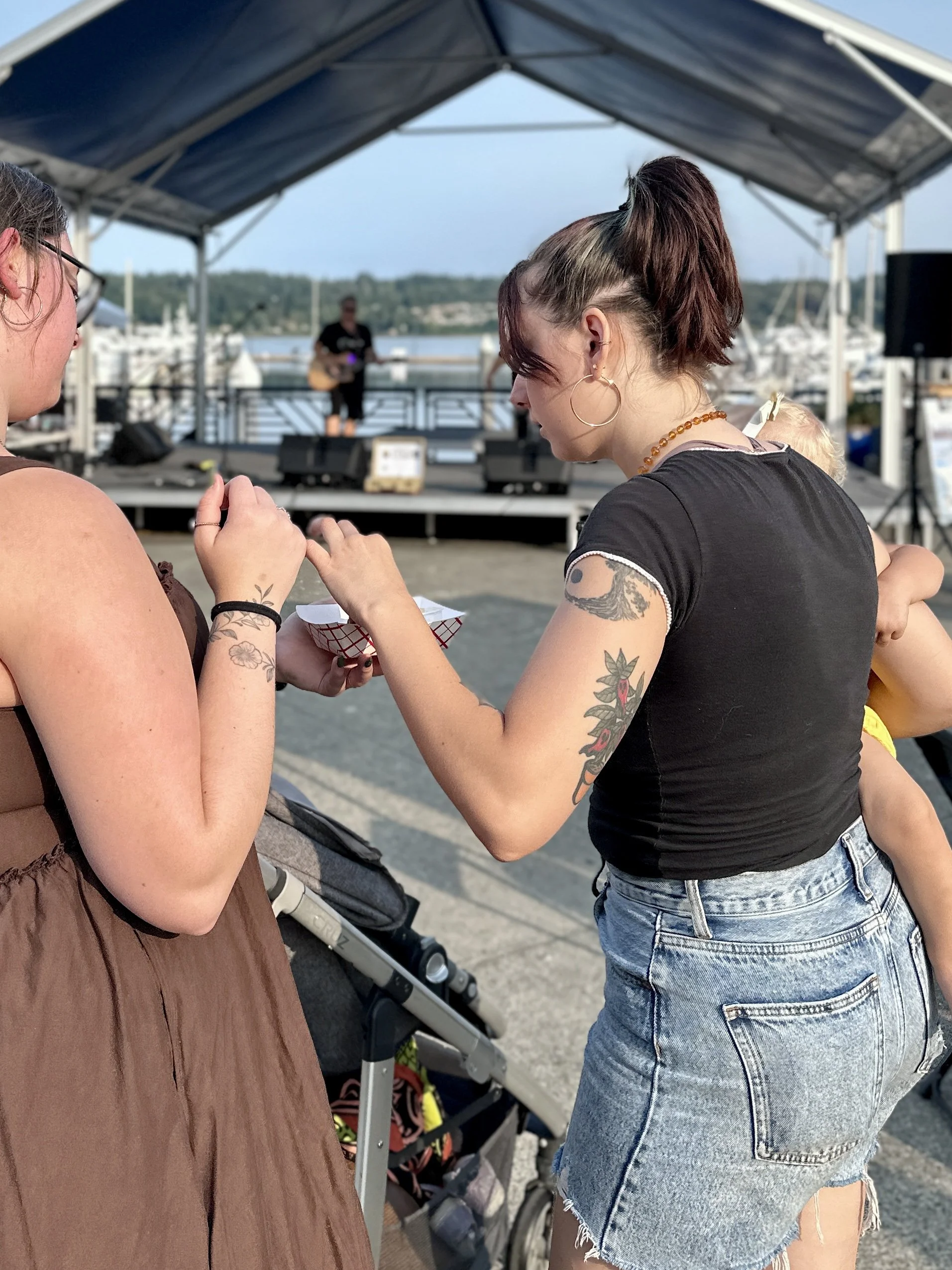 Two young women talking outdoors under a canopy near a marina, with a musician playing guitar on a small stage in the background.