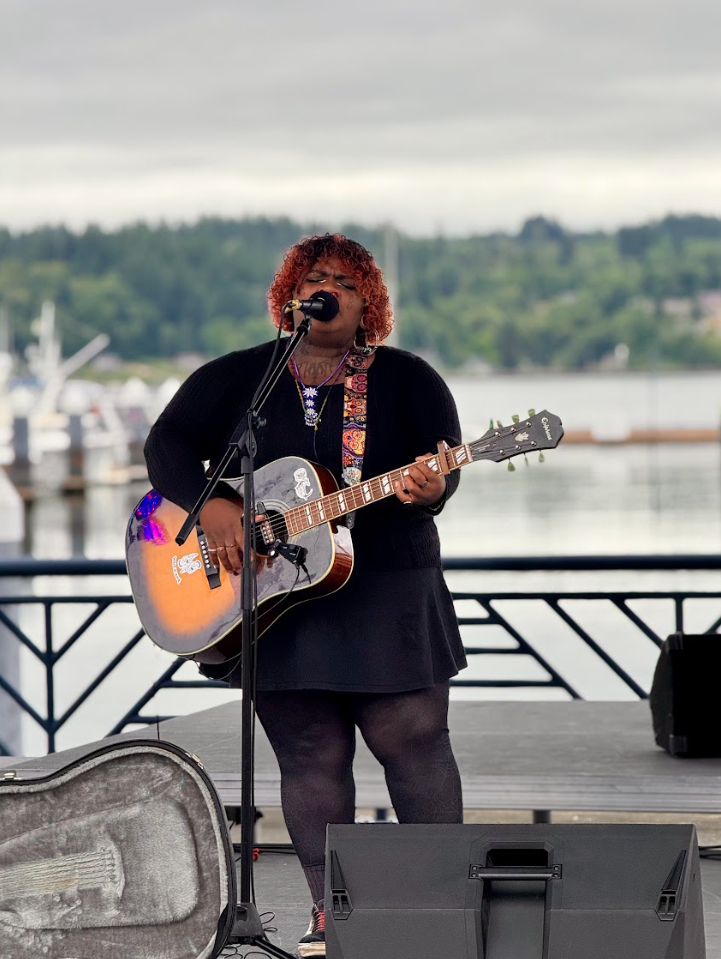 A woman with curly hair singing into a microphone while playing an acoustic guitar on an outdoor stage near a body of water.