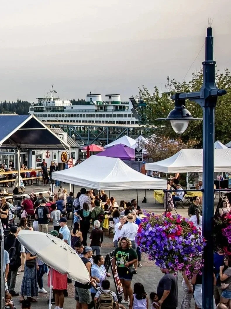 People attending an outdoor market or festival with vendor tents, a large boat in the background, and decorative hanging flower baskets.
