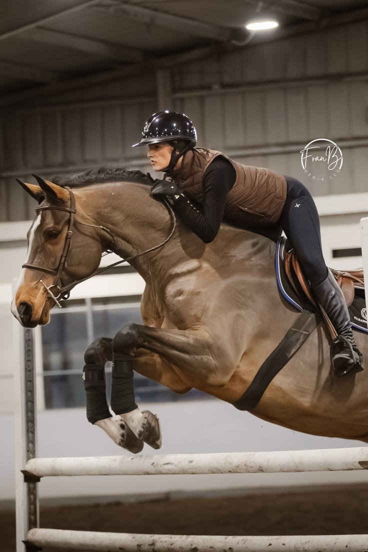 A woman riding a horse over a jump in an indoor equestrian arena.