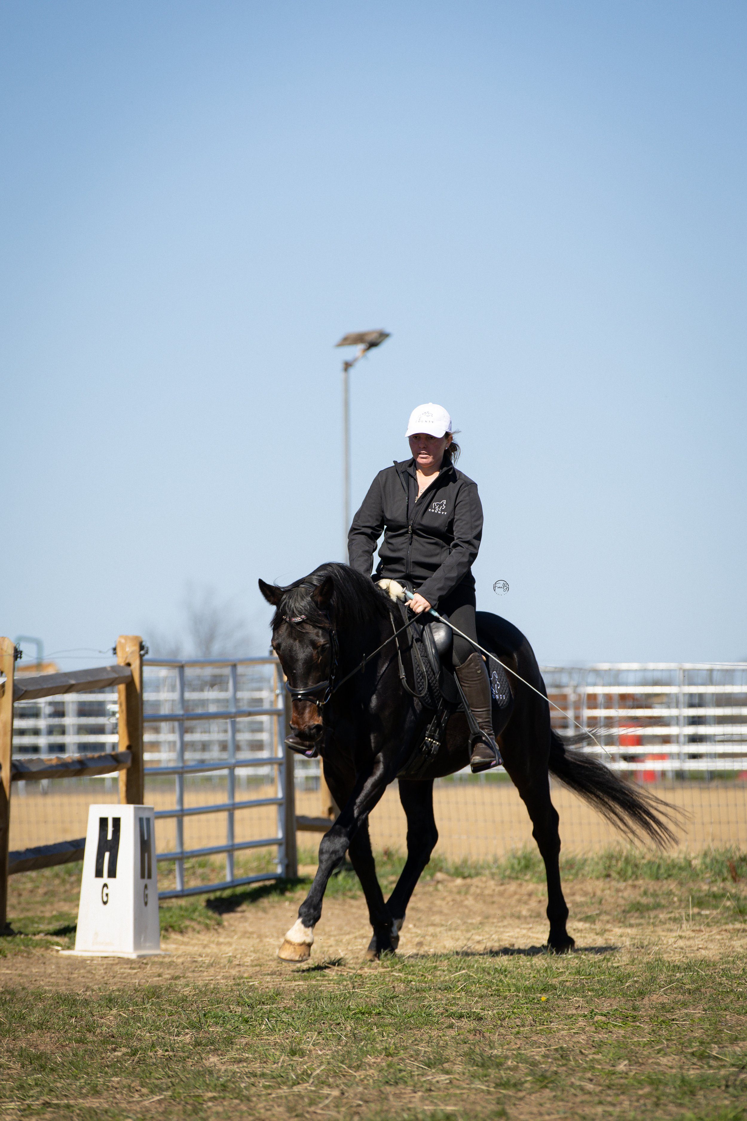 A woman wearing a white cap and black jacket riding a dark horse in an outdoor equestrian arena with fencing, markers, and clear sky.