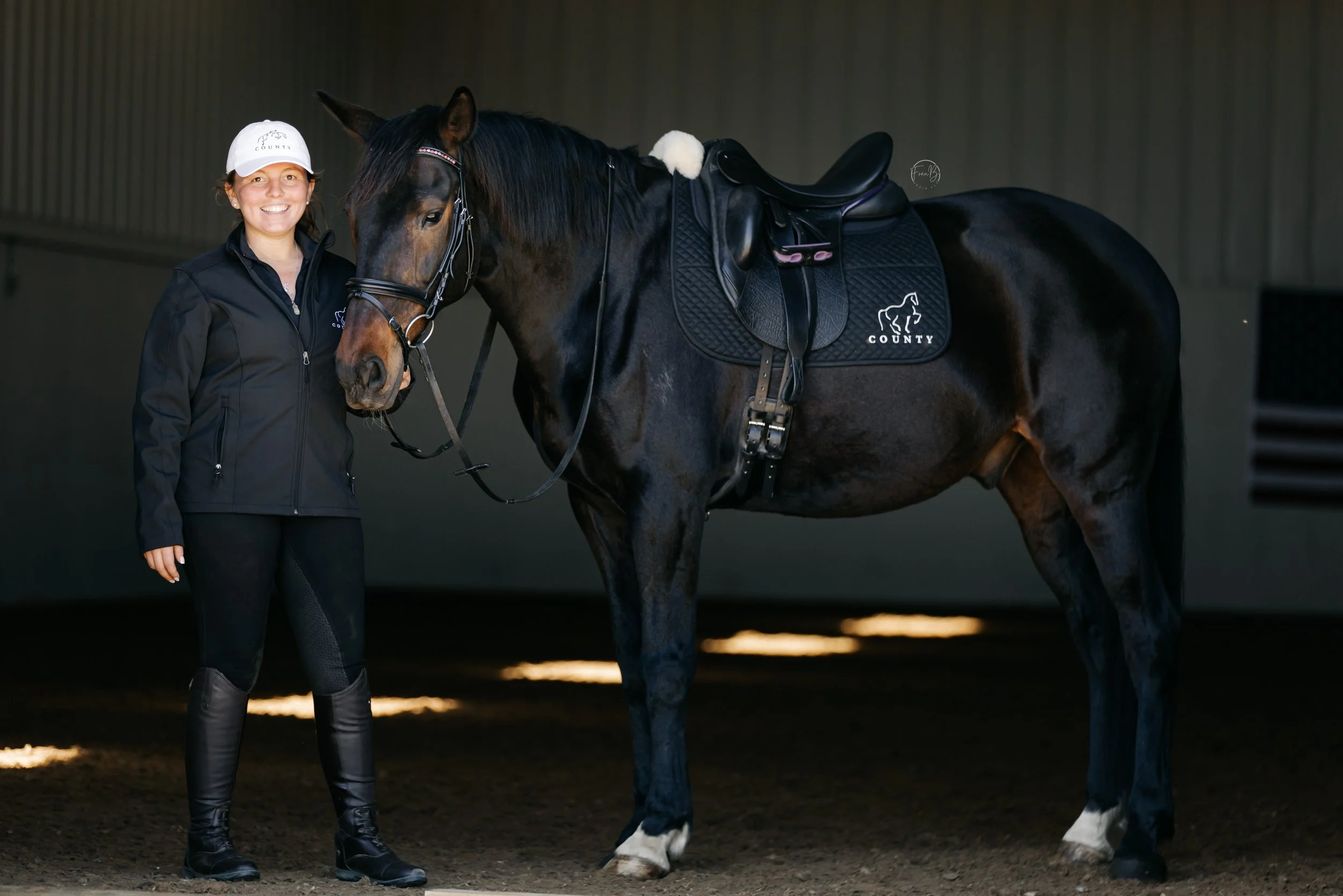 A smiling woman wearing black riding gear and a white cap standing next to a dark brown horse with a shiny coat, inside an indoor riding arena.