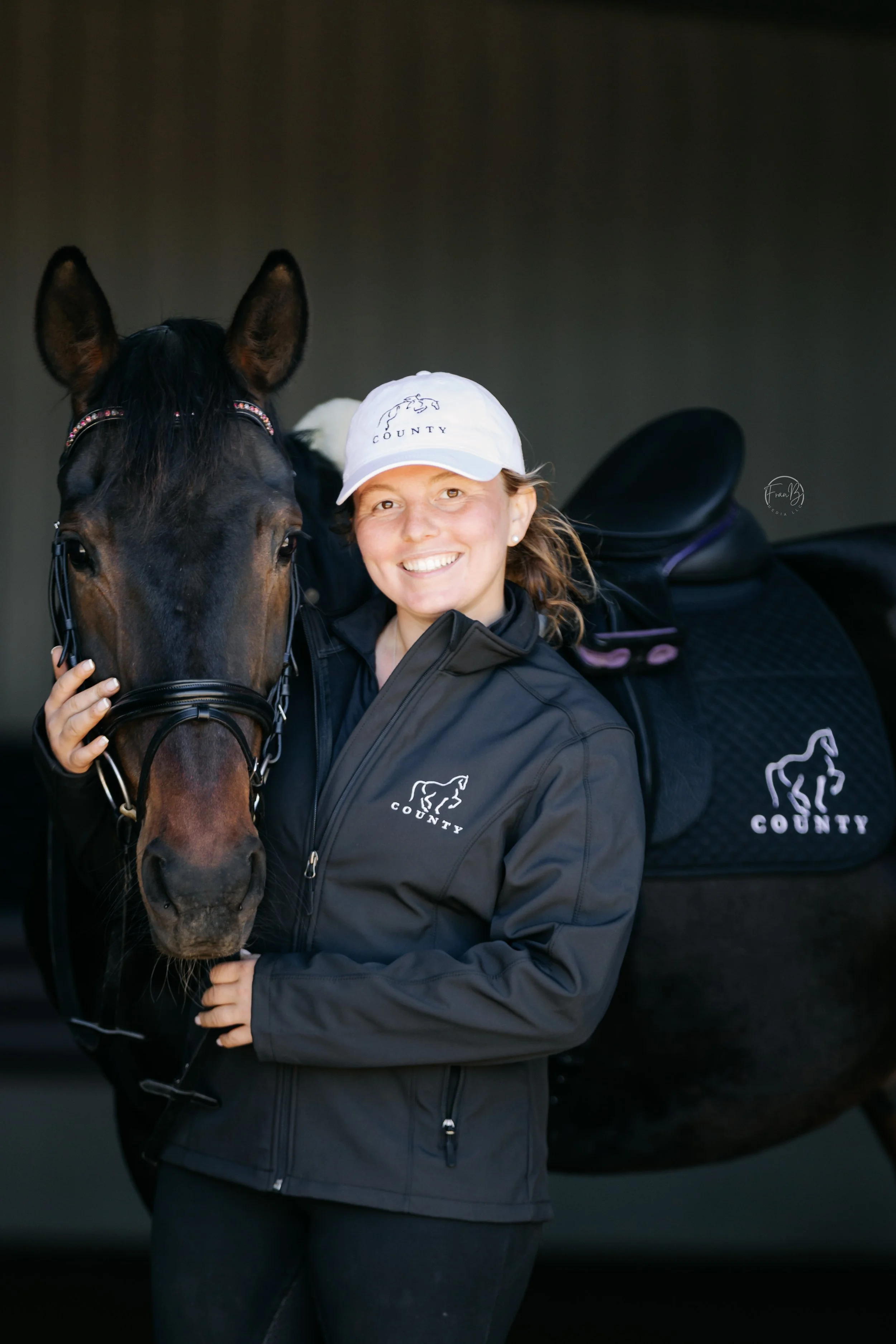 A woman smiling, wearing a white cap and a black jacket with a horse logo, is standing next to her dark brown horse, holding its bridle. The horse's head is close to her face, and she appears happy.