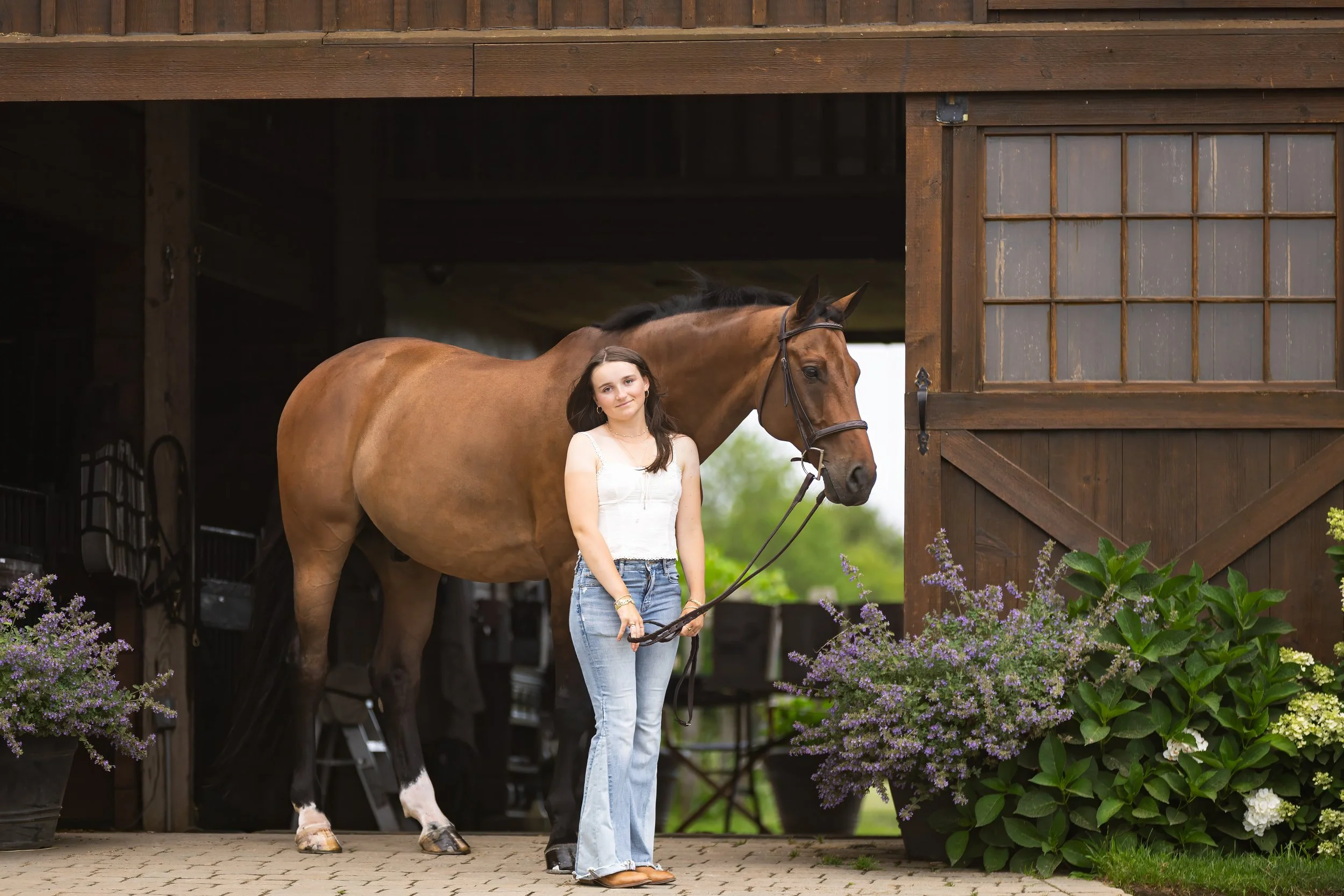 A woman in a white top and jeans holding a brown horse's reins stands outside a wooden stable. The horse is partially visible, with its head near the woman. There are potted flowering plants in the foreground.