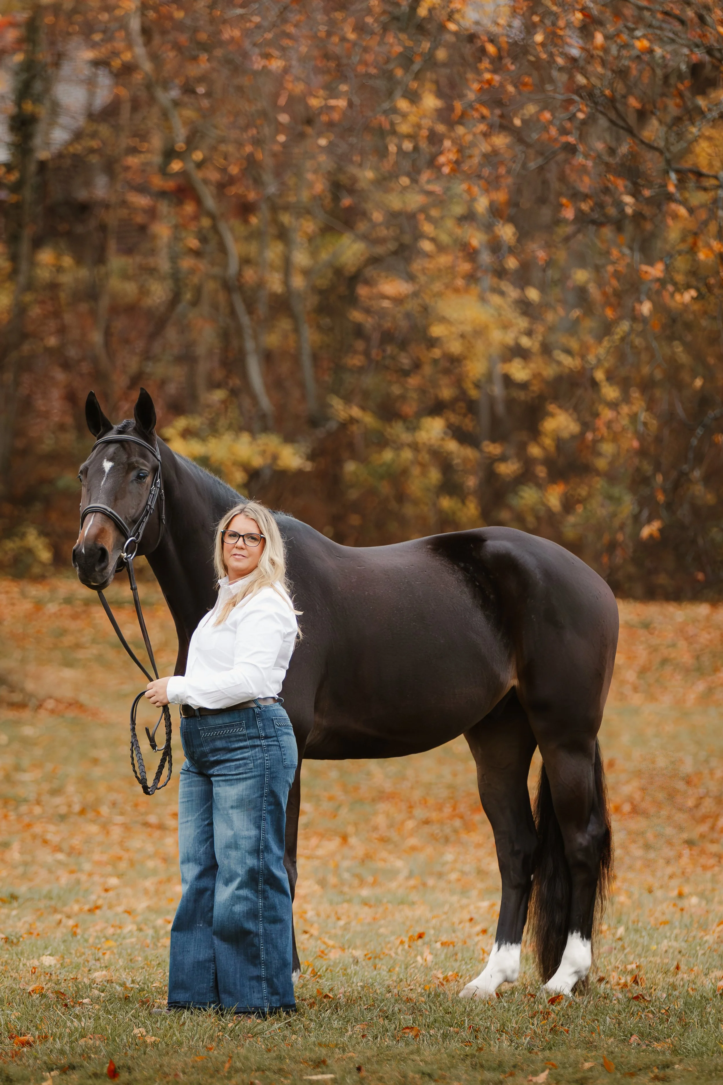 A woman with glasses and long blonde hair wearing a white shirt and jeans standing outdoors holding a black horse with a white star on its forehead. The background features autumn-colored trees with orange and yellow leaves.