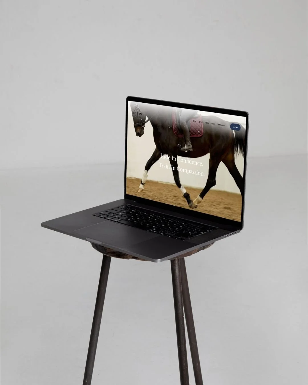 A laptop on a wooden stool displays a website with a horse and a jockey, and the text reads "Ride In Confidence. Train In Compassion."