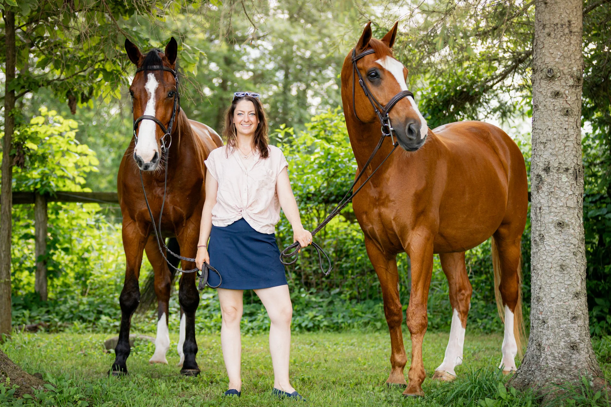 A woman standing outdoors with two brown horses, surrounded by green trees and grass.