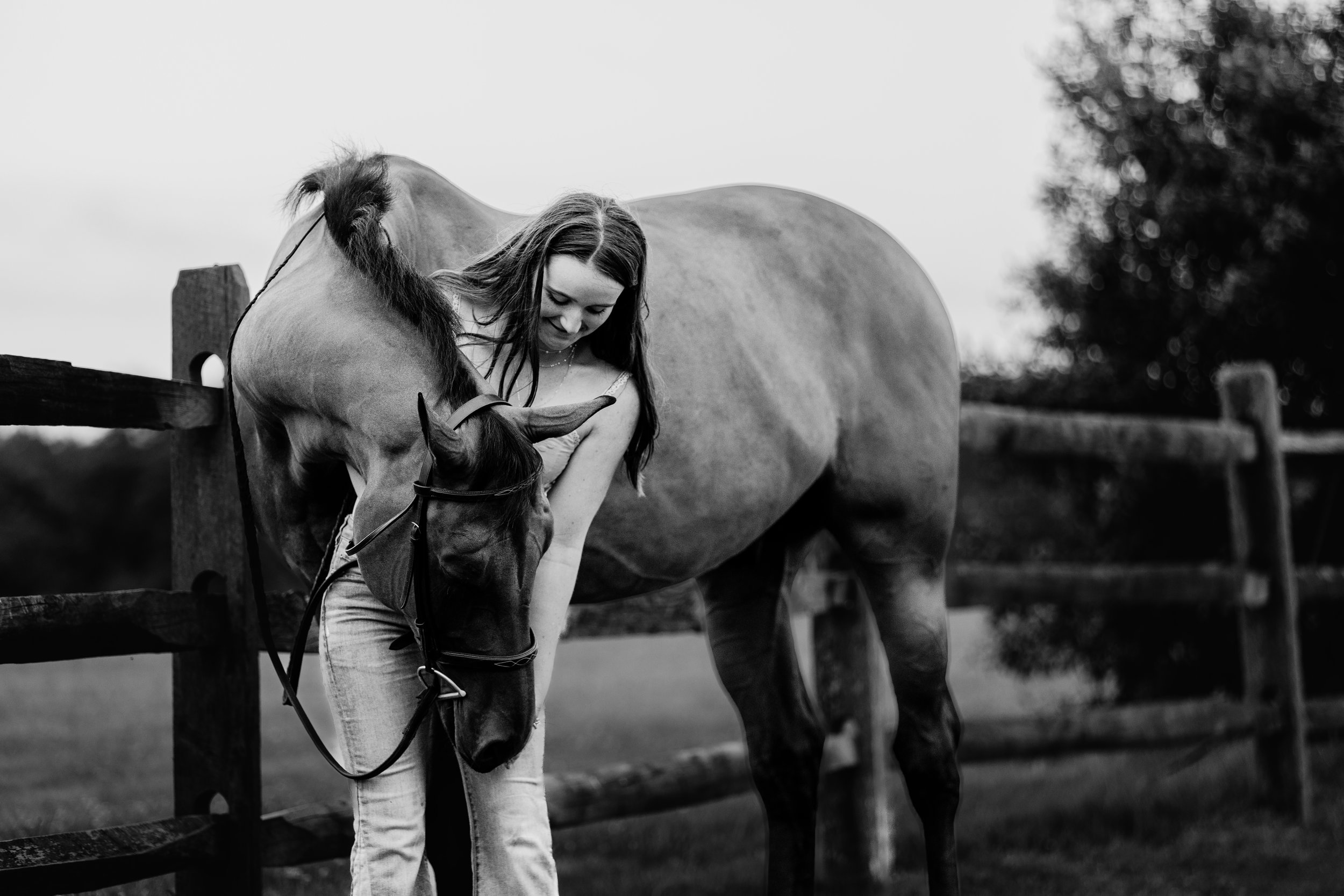 A young woman with long hair and a necklace gently petting a horse beyond a wooden fence in a rural setting, with trees blurred in the background in black and white.