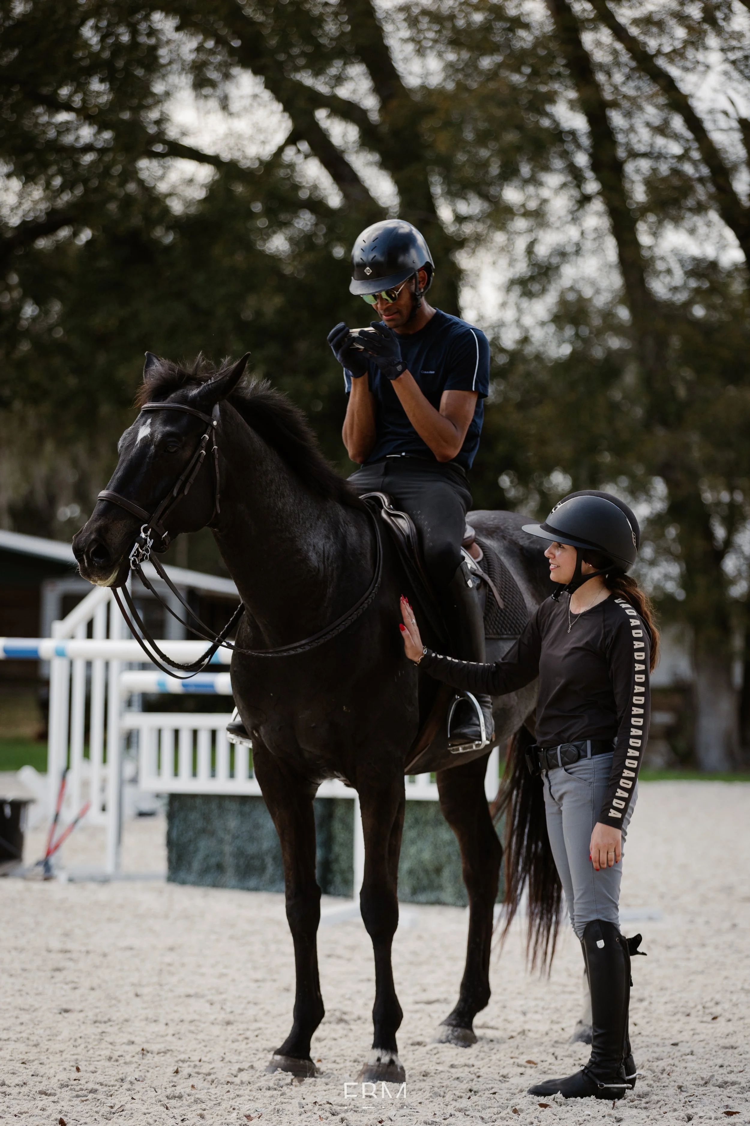 A man in a navy shirt and riding helmet sits on a dark horse, looking at his phone. A woman in riding gear stands beside him, gently holding the horse's neck, within a riding arena with jumps and trees in the background.
