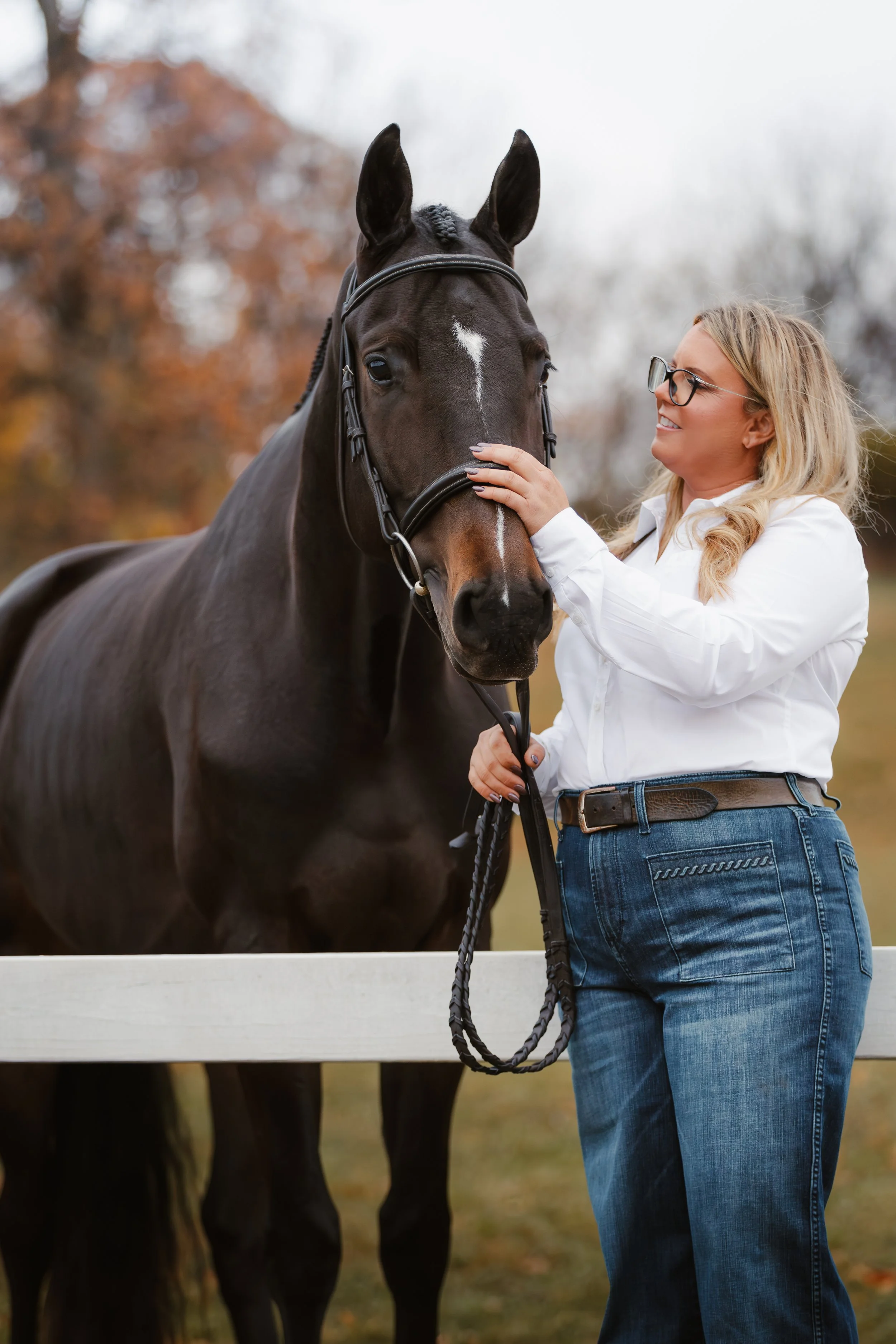 A woman with blonde hair, glasses, and a white shirt is holding the bridle of a dark bay horse with a white star-shaped marking on its forehead, standing next to a fence outside in autumn.