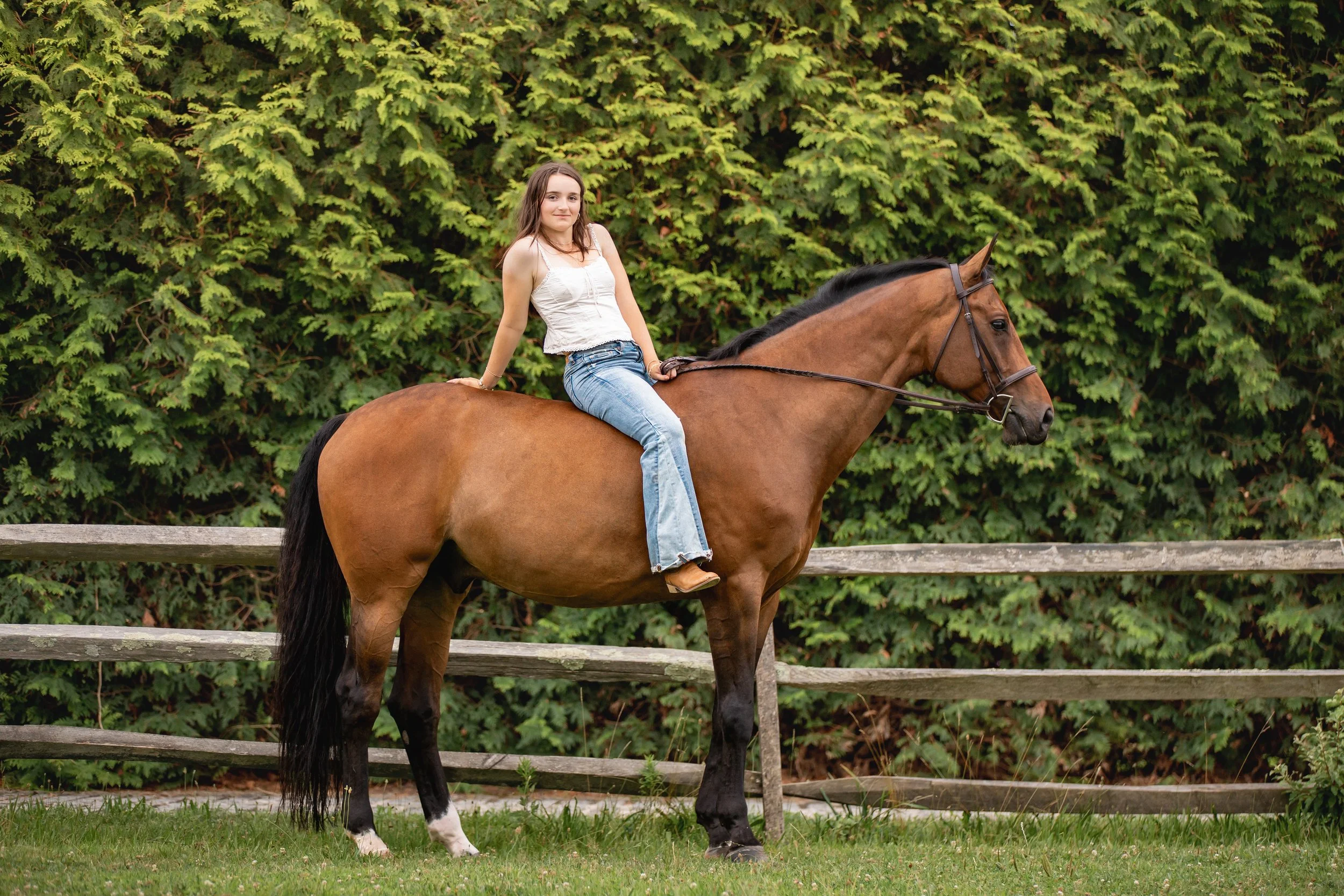 A young woman sitting on a brown horse with black mane, in a green outdoor setting, with a leafy background and wooden fence.