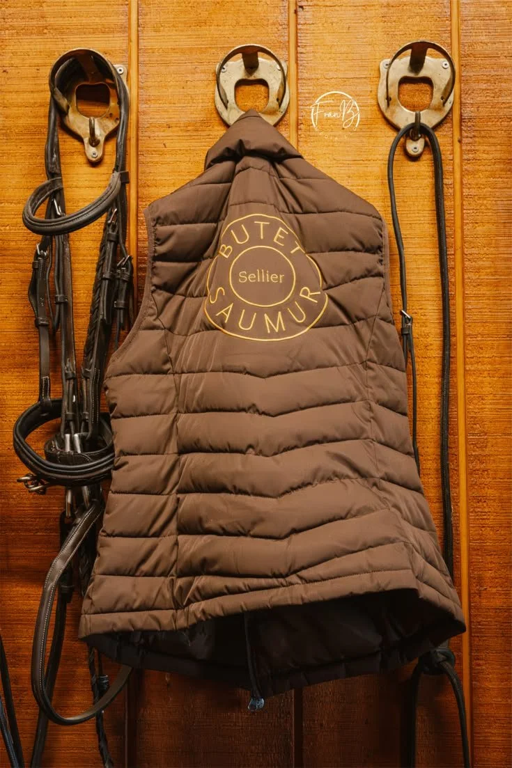 A quilted brown jacket hanging on a wood wall, with horse tack and stirrups on either side.