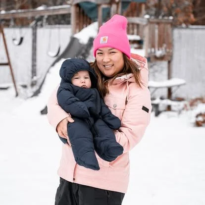 Frances in a pink jacket and pink beanie holding a baby in winter clothes outside in the snow.