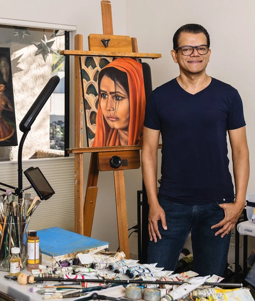 An artist stands next to a portrait of a woman with a serious expression, wearing a red headscarf. The artist is wearing glasses and a navy blue shirt, smiling slightly, with art supplies and unfinished paintings around him in the studio.