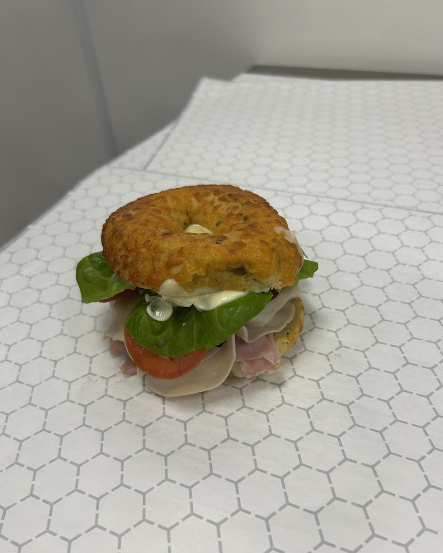 An open-faced bagel sandwich with lettuce, tomato, turkey, ham, and mayonnaise on a white hexagonal-patterned paper surface.