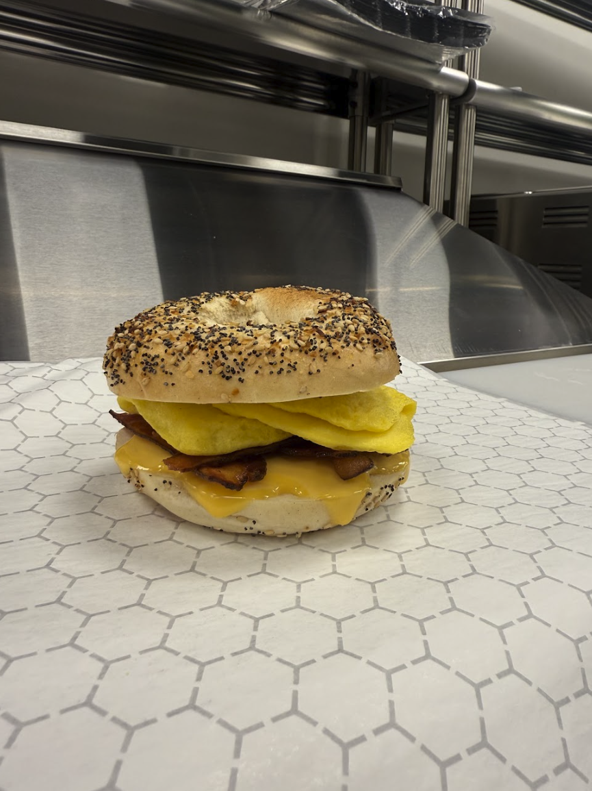 Bagel sandwich with cheese, bacon, and egg on honeycomb-patterned paper in a commercial kitchen.