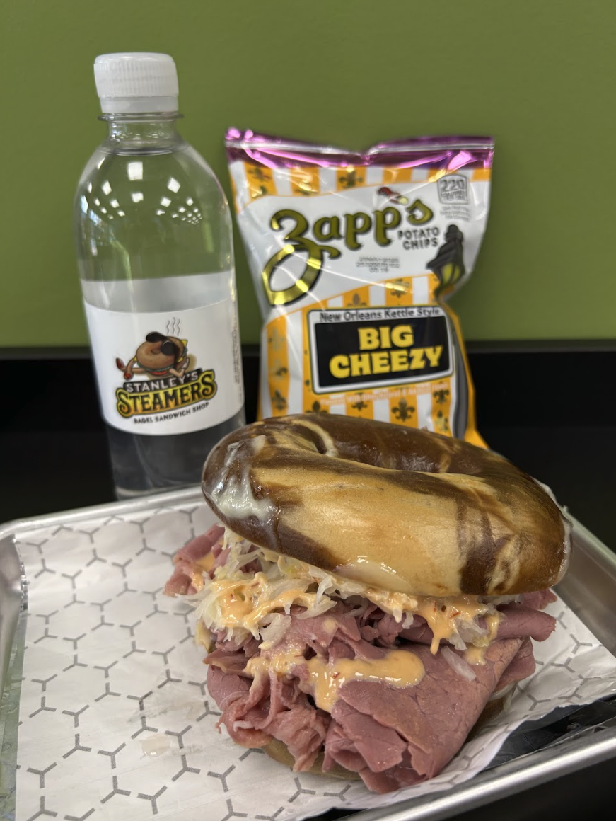 A roast beef sandwich with shredded meat, cheese, and sauce in a hoagie bun, served on a tray with a bag of Zapp's Big Cheezy potato chips, a bottle of water labeled Stanley's Steamers, and a green background.