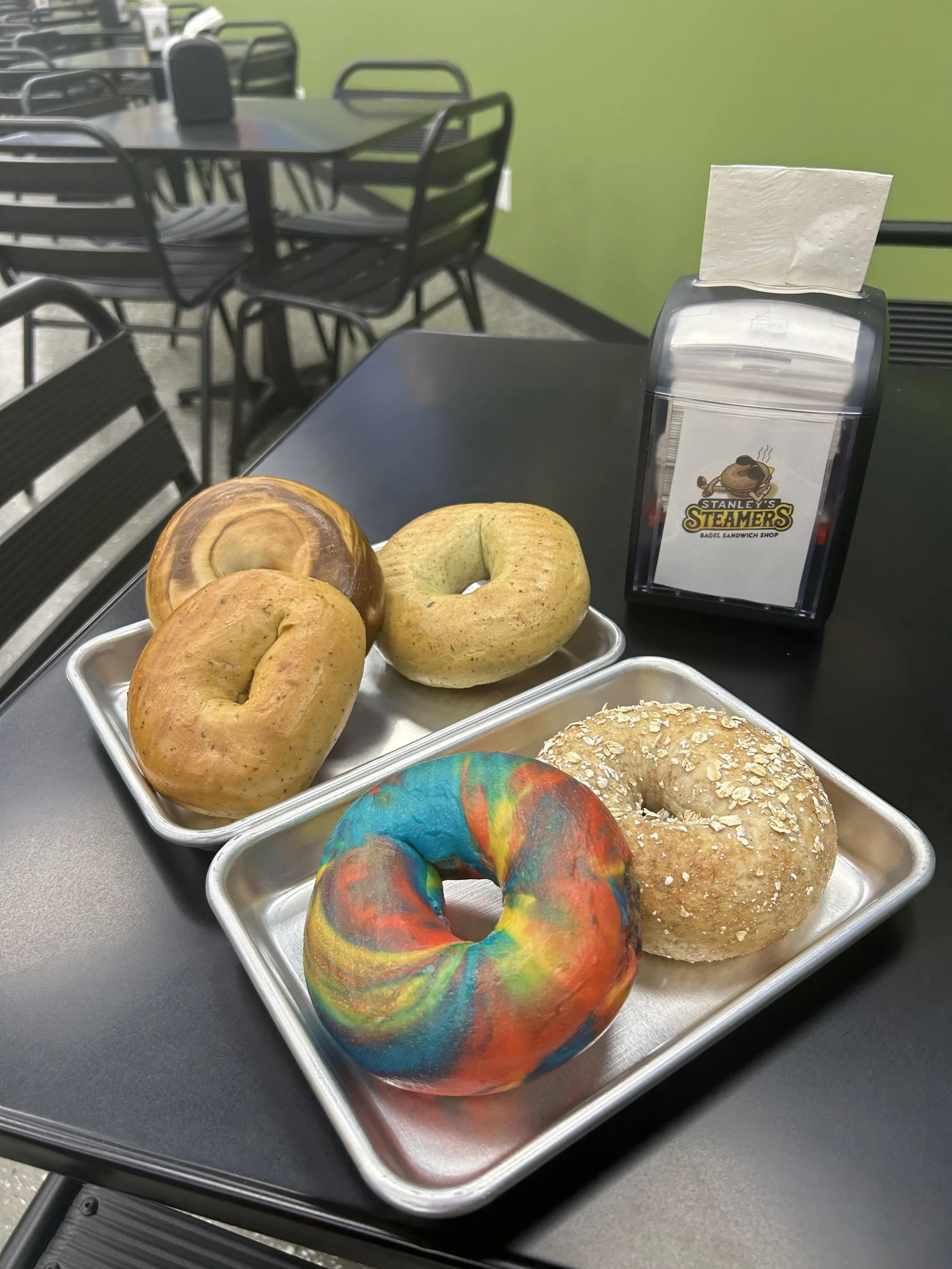 Five bagels on silver trays; colorful rainbow bagel, everything, cinnamon raisin, plain, and oat flakes. A napkin dispenser with a logo for Stanley's Steamers Bagel Sandwich Shop, on a black table in an outdoor seating area with green walls and black metal chairs.