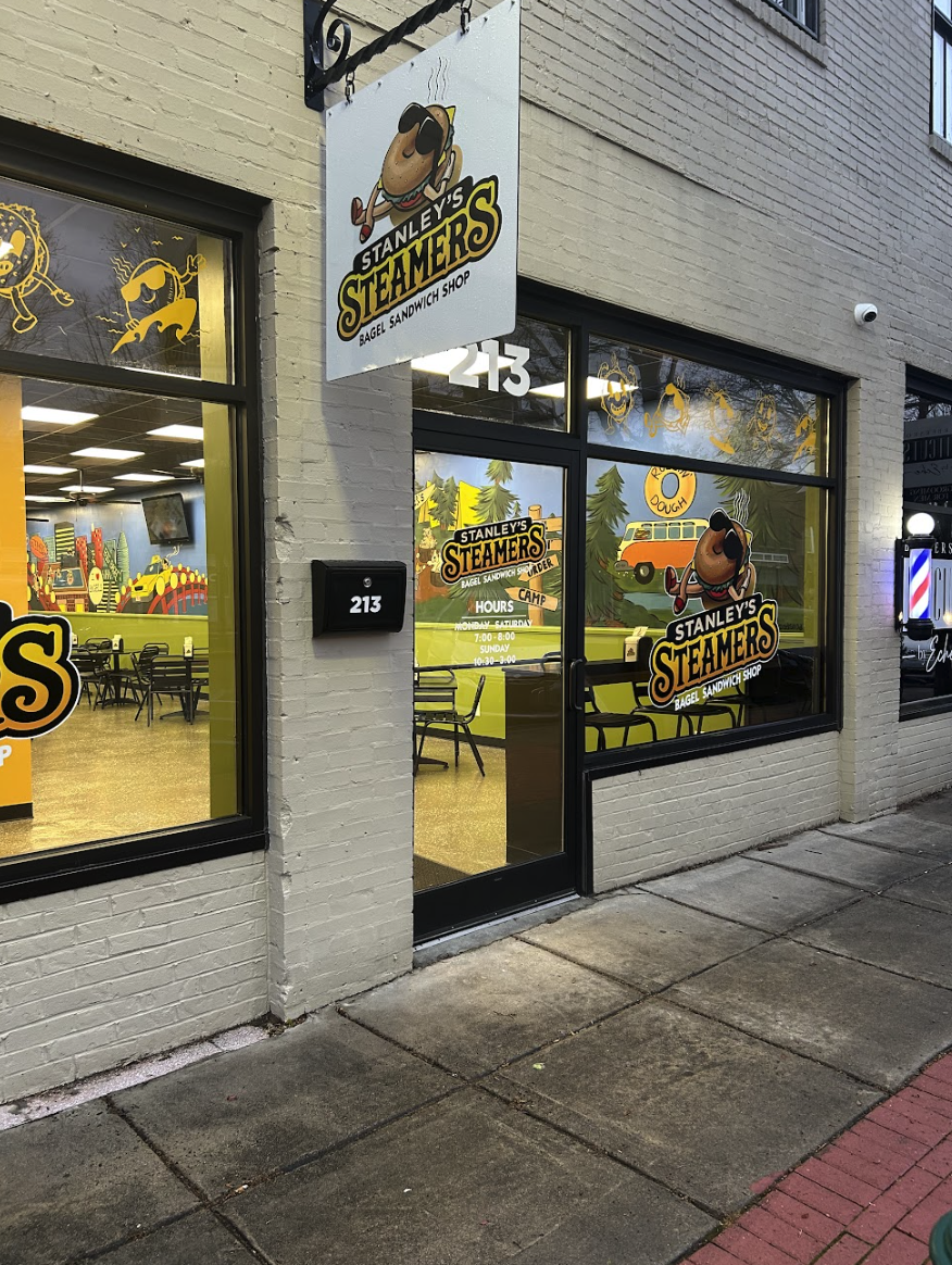 Exterior of Stanley's Steamers bagel sandwich shop with signage featuring a cartoon bagel character wearing sunglasses and headphones. The shop has large front windows with yellow designs, and a neighborhood sidewalk in front.