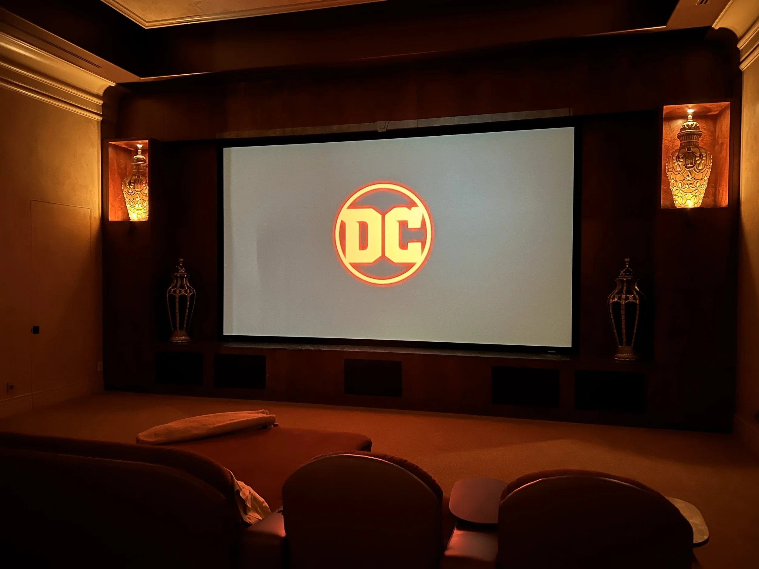 Home theater room with a large screen showing the DC logo, beige sofa, and decorative lamps on the wall.
