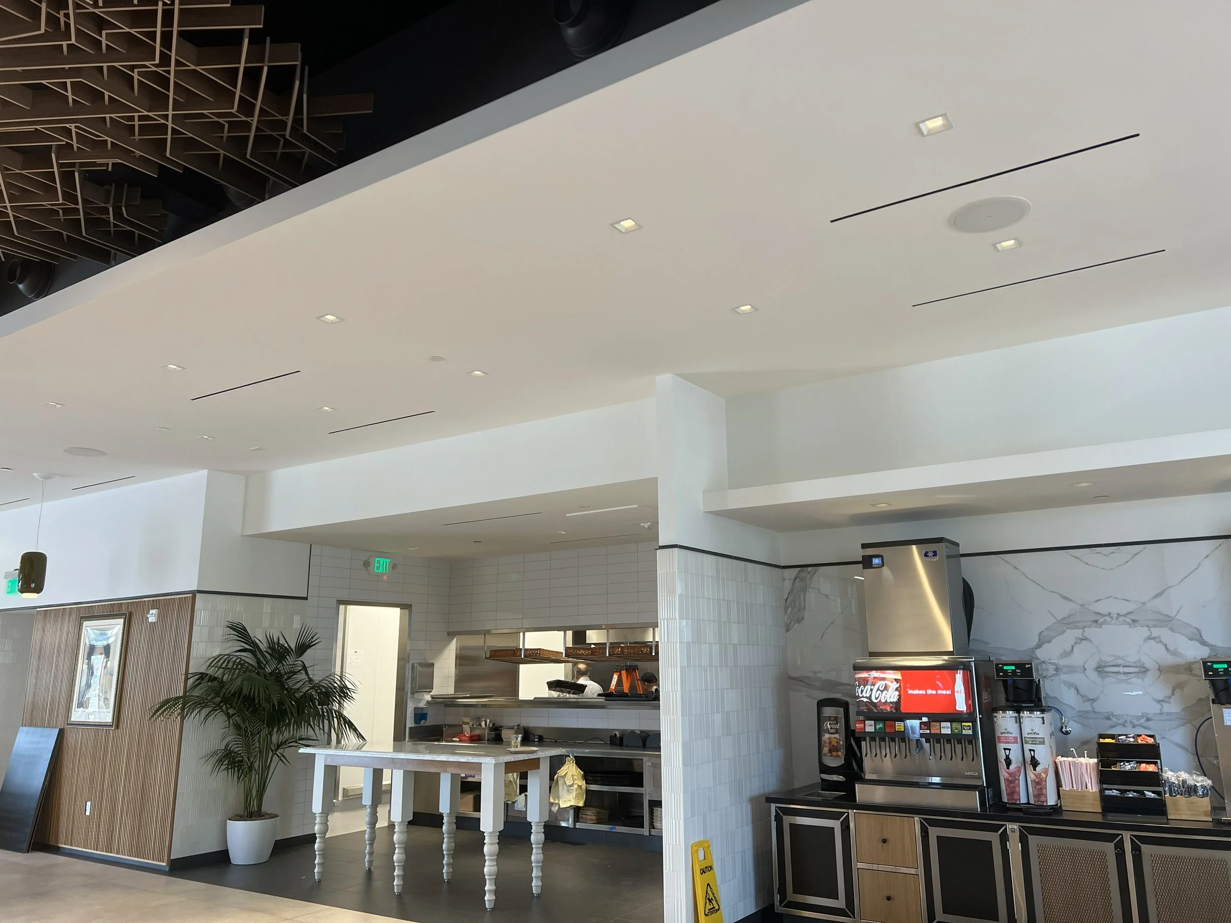 Interior view of a modern cafeteria with a soda fountain, snack station, a potted plant, and a visible kitchen area in the background.