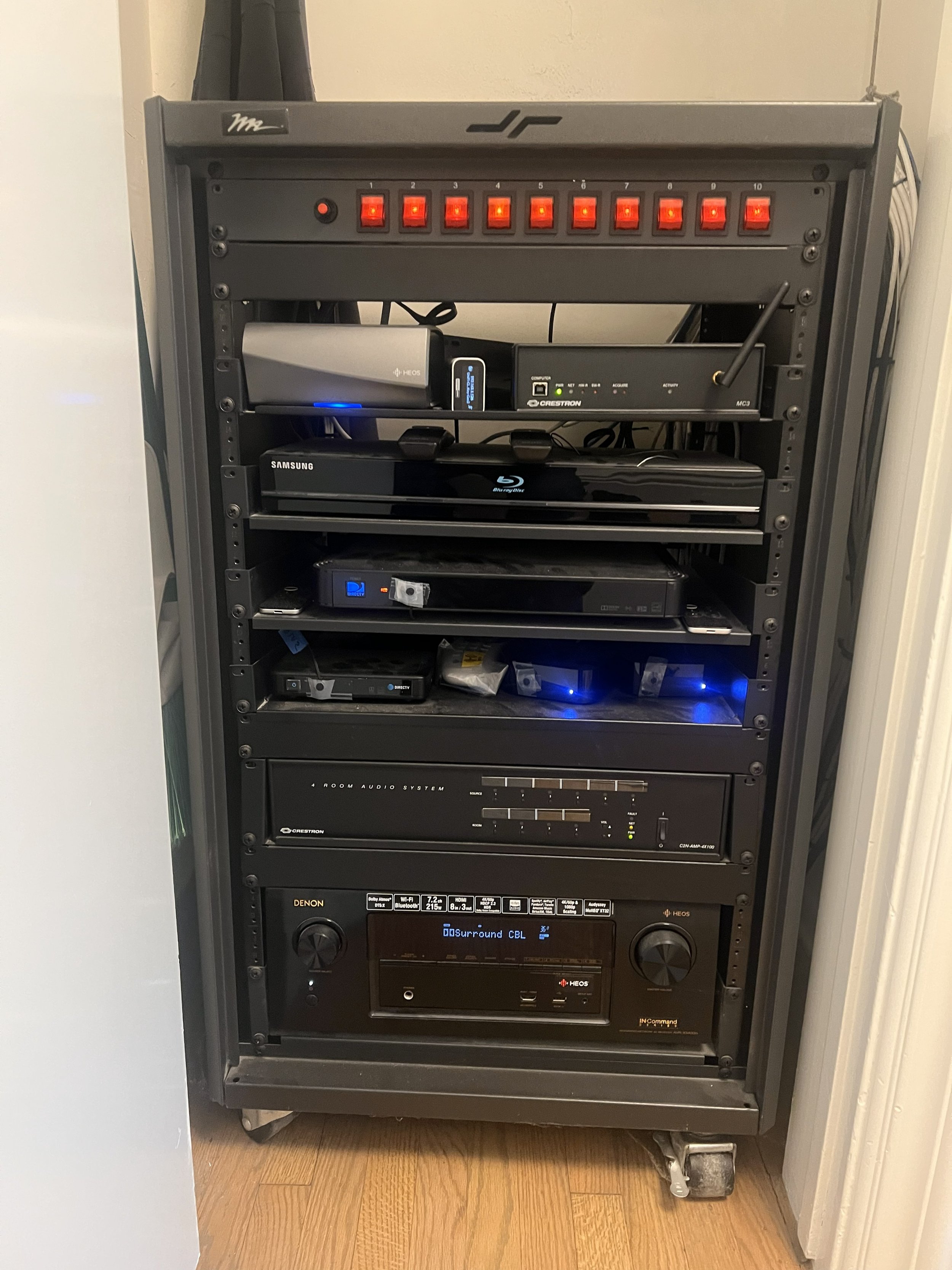 A rack of electronic equipment including a power strip with multiple switches on top, a Blu-ray player, an audio receiver, and other media devices, all stacked on metal shelves on a wooden floor for video system.