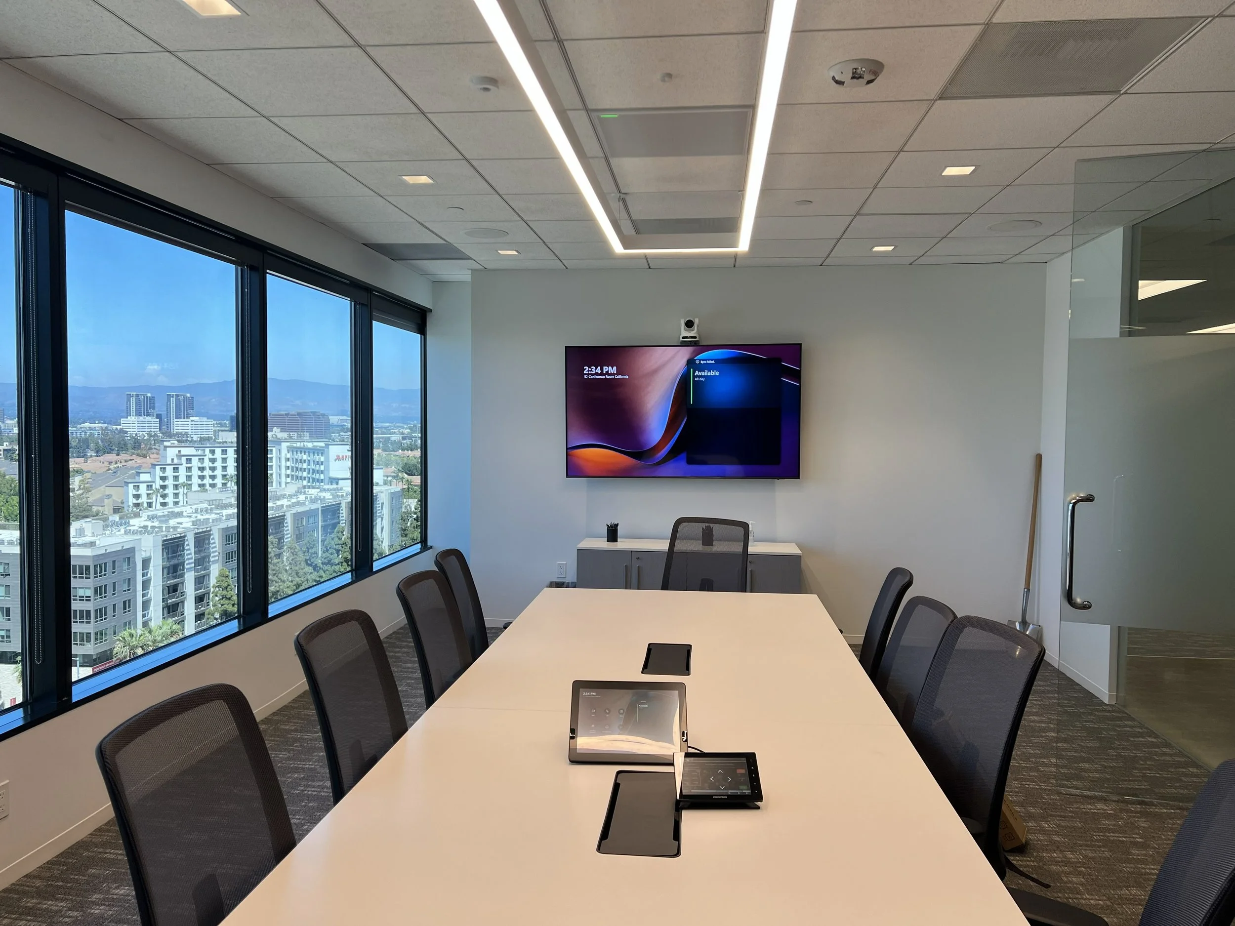 Empty modern conference room with a large white table, black mesh chairs, a wall-mounted TV, conference phone, and a view of city buildings through large windows.