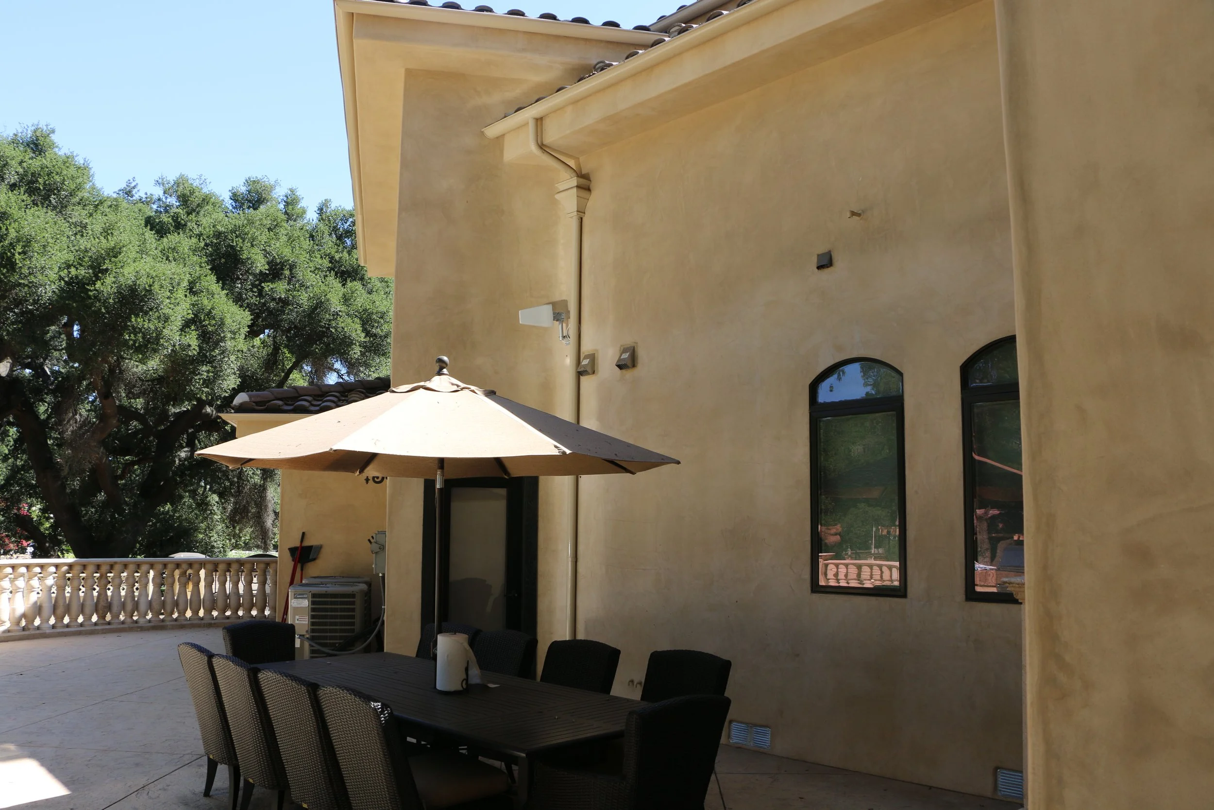 Outdoor patio area with a rectangular table, six chairs, and a large beige umbrella, set against a yellow stucco wall with two arched windows, trees in the background, and an air conditioning unit.