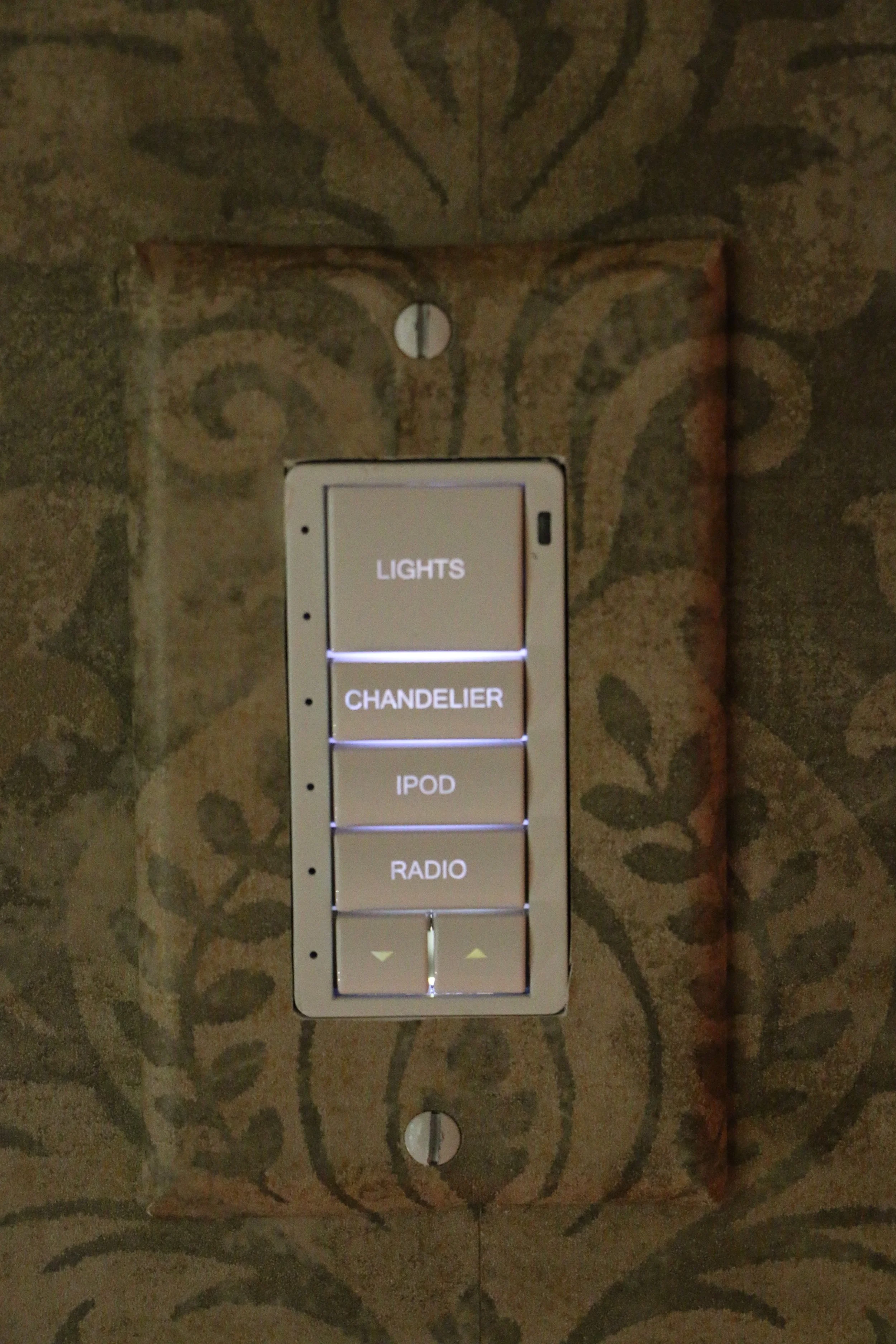 A wall-mounted control panel with illuminated buttons labeled LIGHTS, CHANDELIER, IPOD, RADIO, and arrows for volume adjustment. 