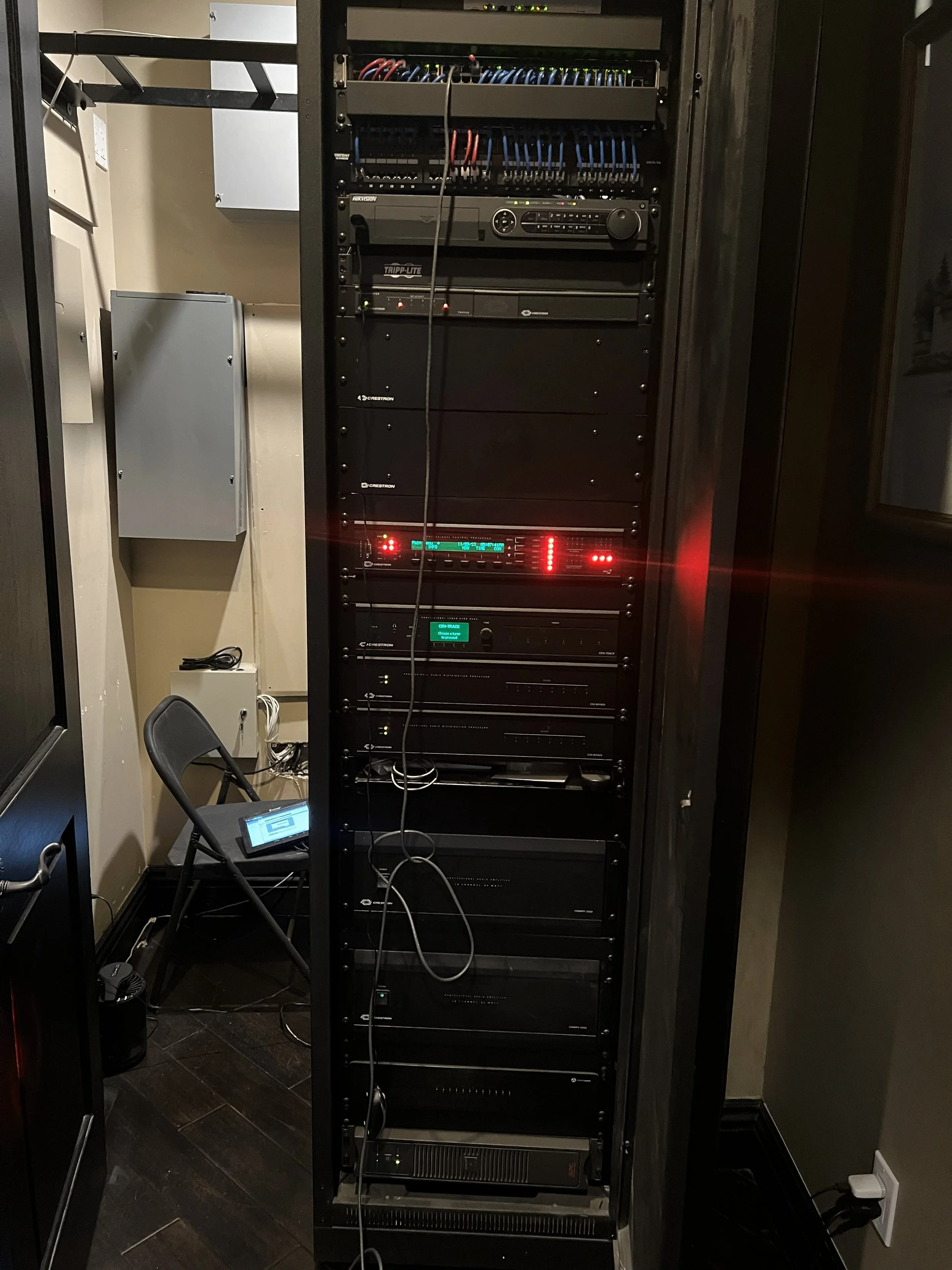A server rack filled with network and audio equipment, located in a room with a chair and a monitor on the floor.