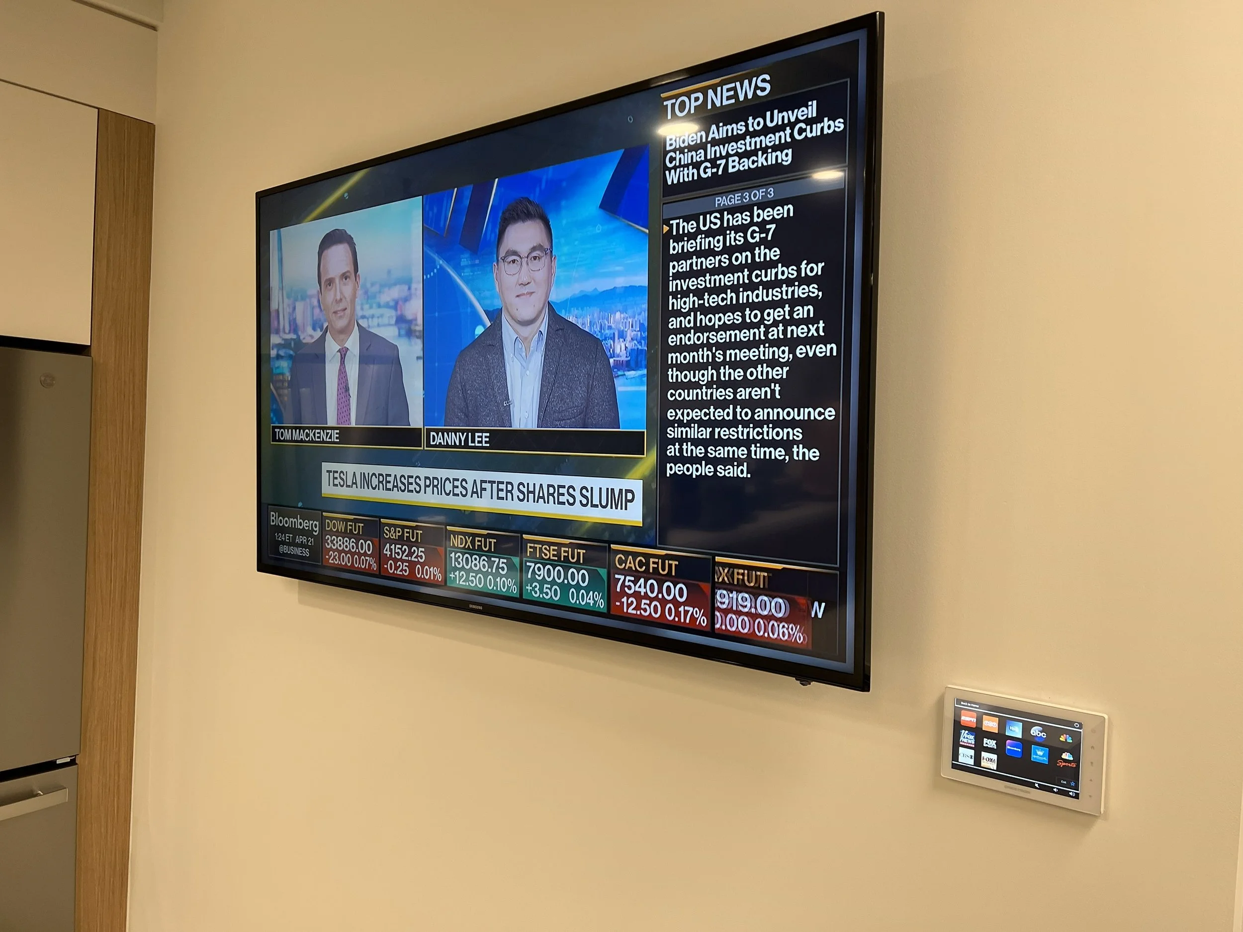 Television screen displaying financial news with two male anchors, Tom Mackenzie and Danny Lee, and stock market data at the bottom, mounted on a beige wall with a control panel nearby.