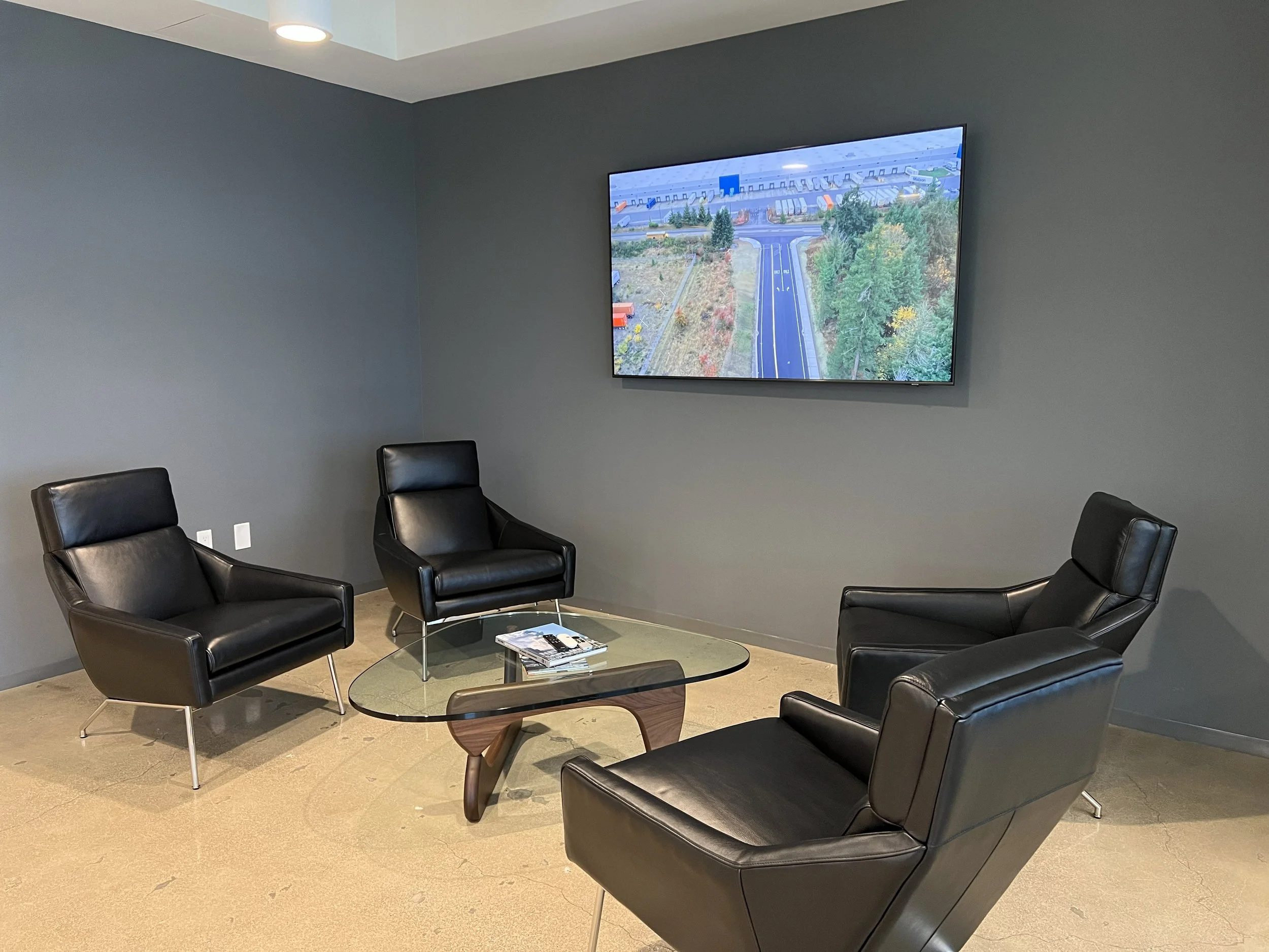 A modern conference room with four black leather armchairs arranged around a glass coffee table. A large flat-screen TV mounted on a gray wall displays an aerial view of a road and surrounding landscape.