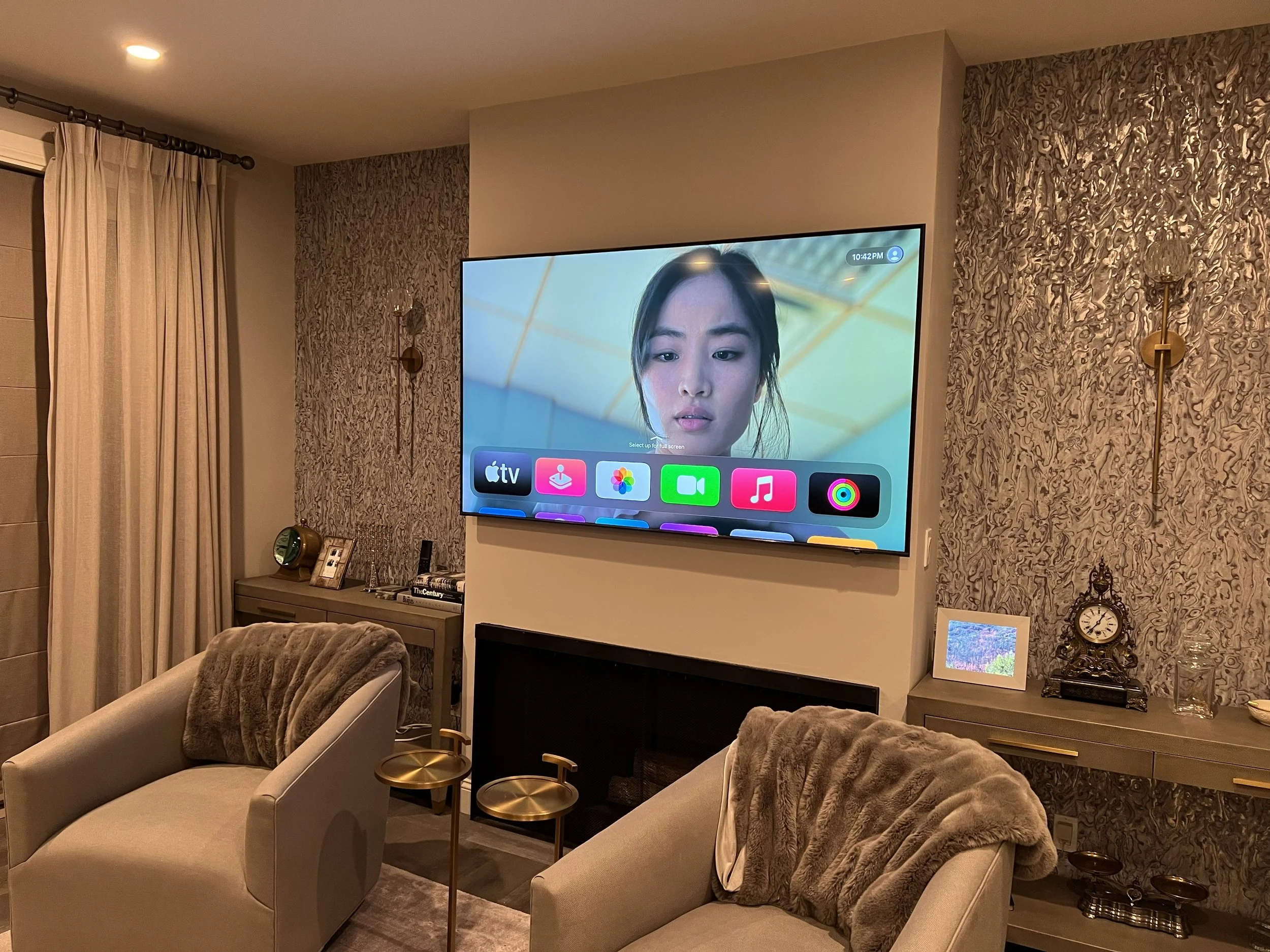 Living room with beige armchairs, side tables, wall-mounted TV displaying a woman's face in a video call, fireplace, patterned wallpaper, clock, framed photo, and decorative items.