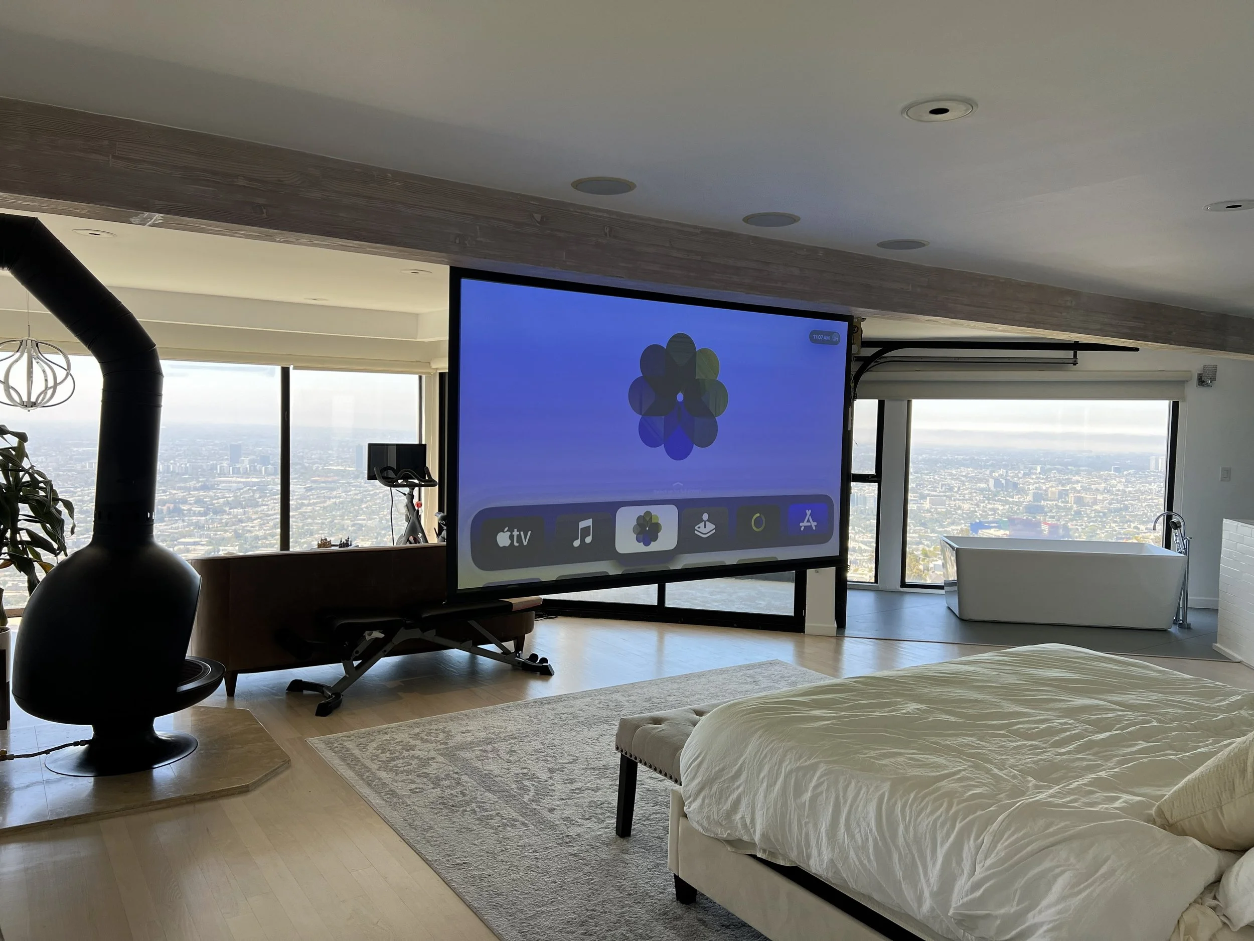 Modern living room with large windows showing city view, a bed with white bedding, a large screen displaying a colorful app, a black wood stove, a bench, a tub near the window, and various home decor elements.