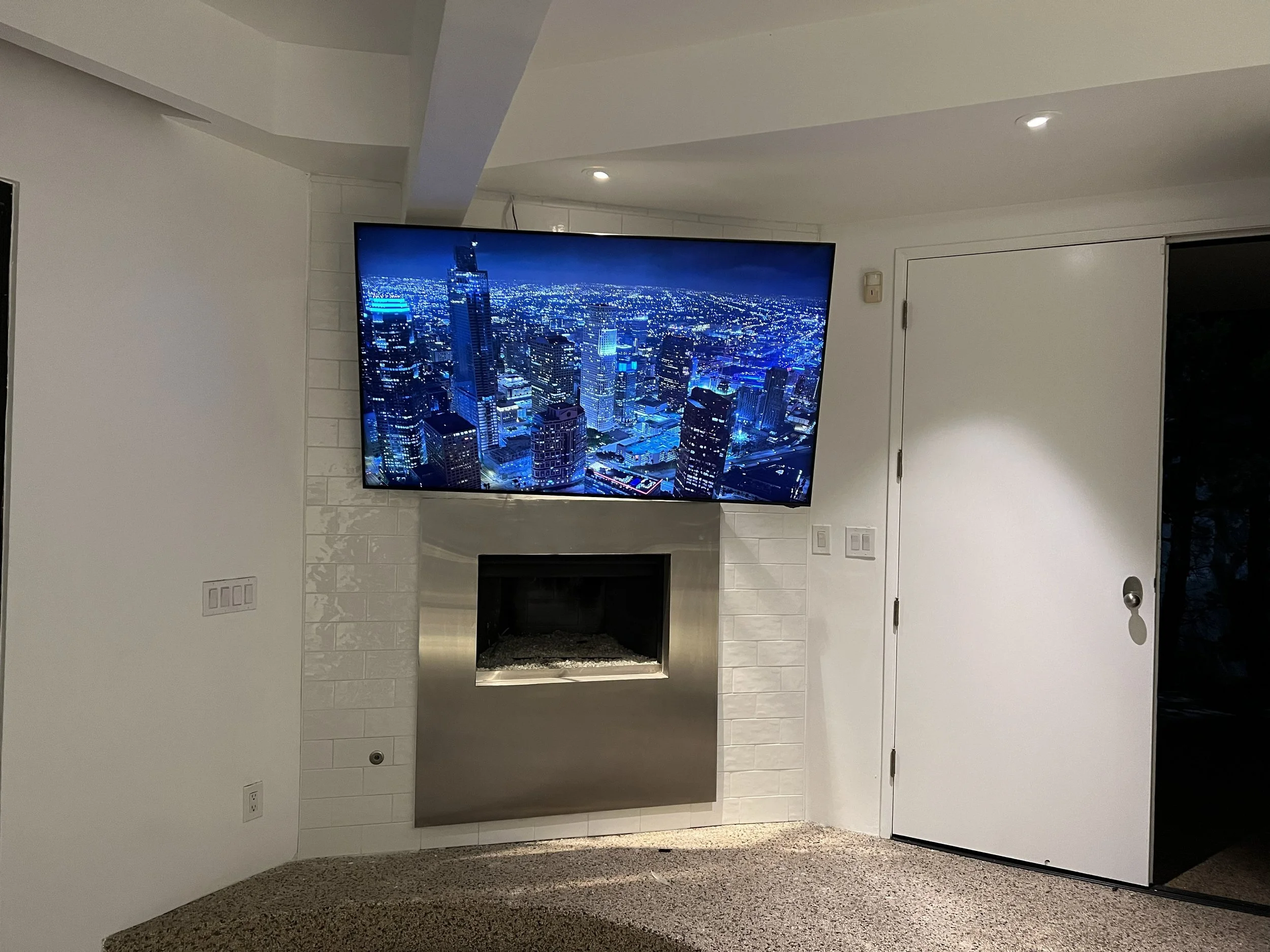 A living room corner with a mounted flat-screen TV displaying a cityscape at night, positioned over a modern fireplace, with a white wall and a white door nearby.