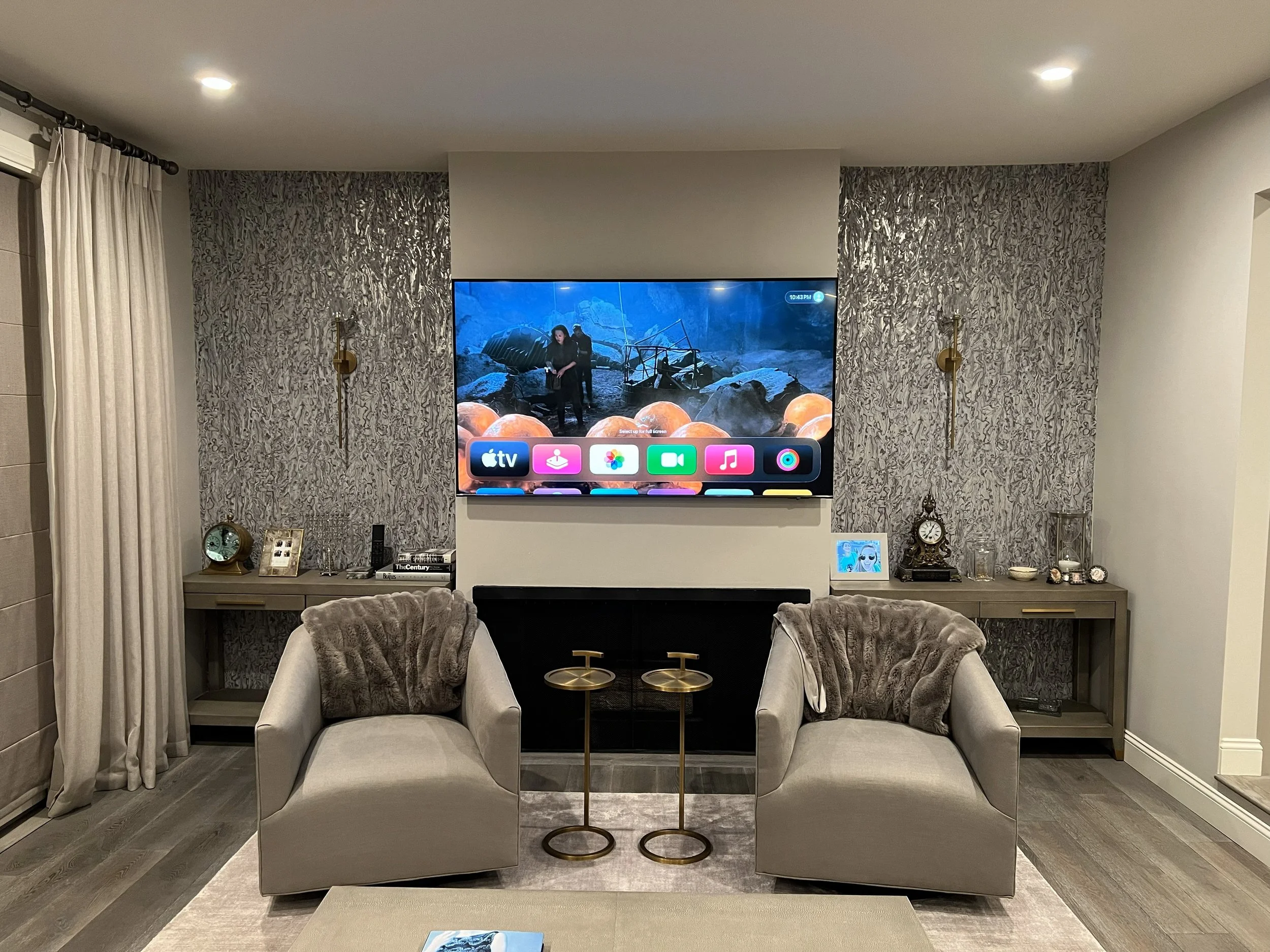 Living room with two beige armchairs with faux fur throws, a wall-mounted flat-screen TV showing a scene from a movie or show, side tables, decorative items, and a patterned accent wall.
