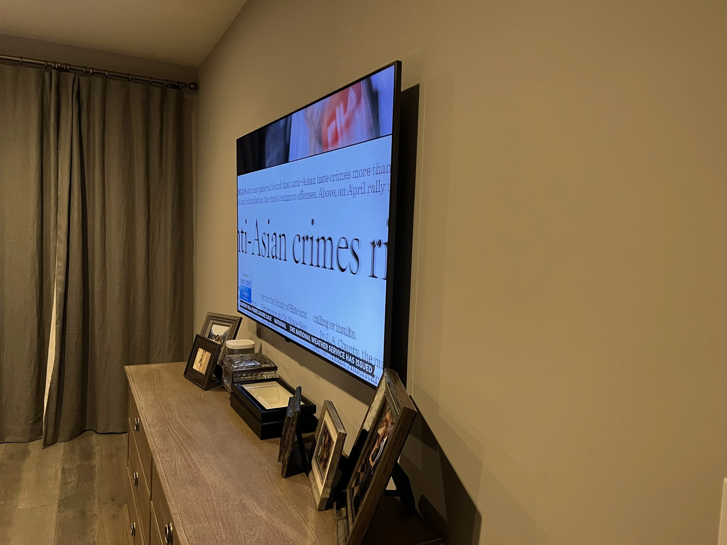 Living room with a wall-mounted TV displaying news, a wooden console table with framed photographs, jewelry box, and decorative items, beige curtains on a window.