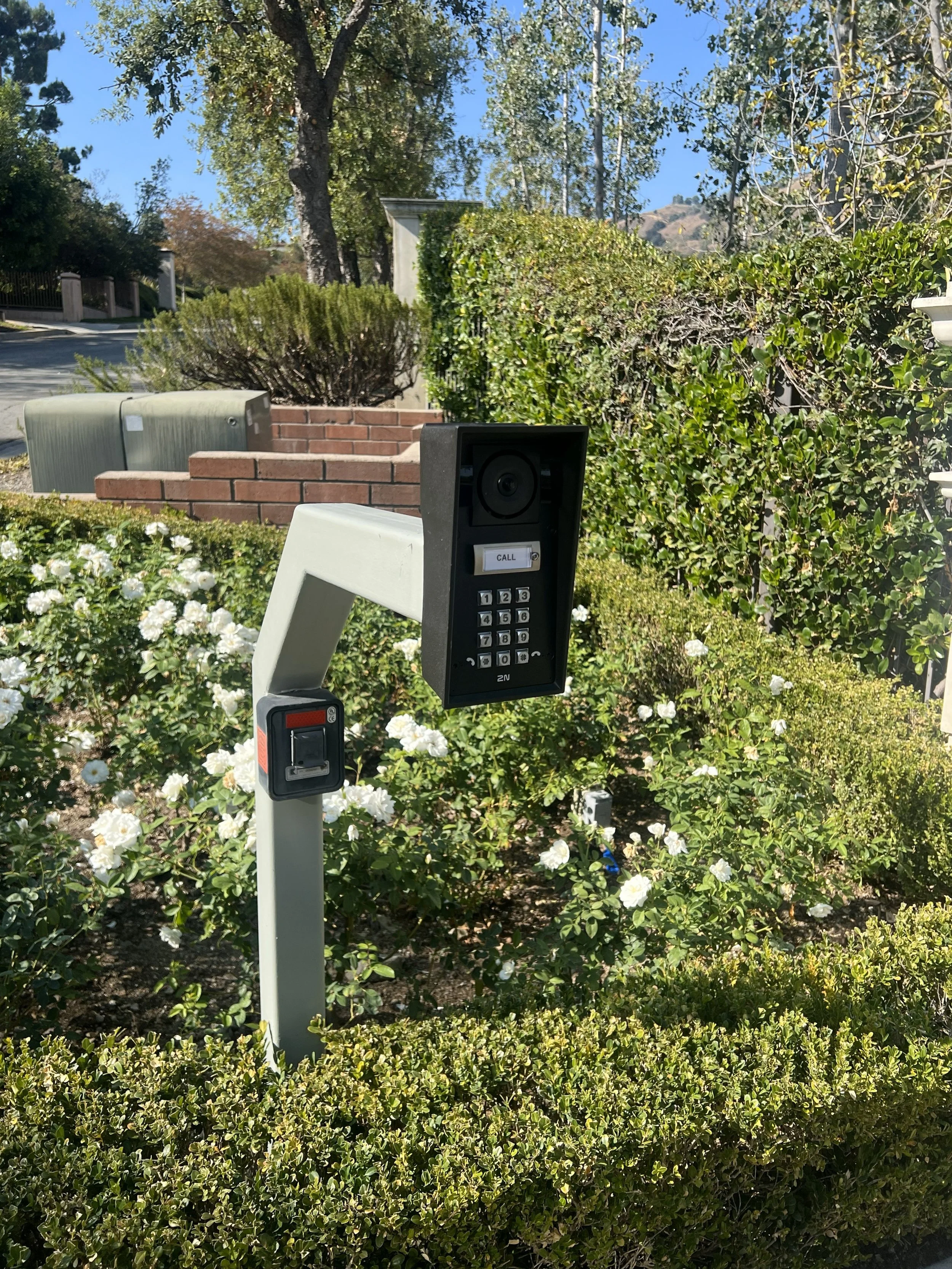 A building intercom system with a camera, call button, keypad, and card reader, installed outdoors among bushes and flowers.