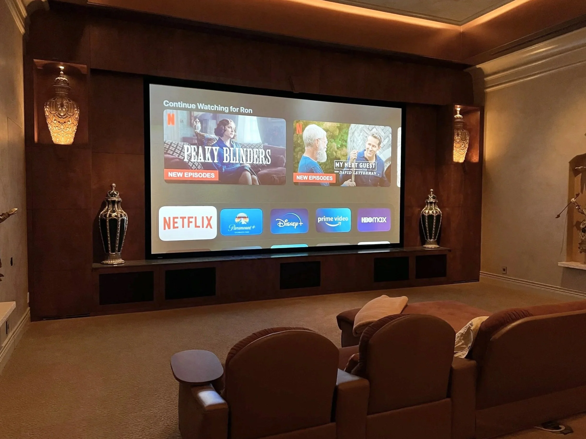 A home theater room with a large screen displaying Netflix's interface, featuring shows 'Peaky Blinders' and 'My Next Guest'. The room has beige carpet, three brown recliner chairs, decorative lamps on either side of the screen, and a small side table.