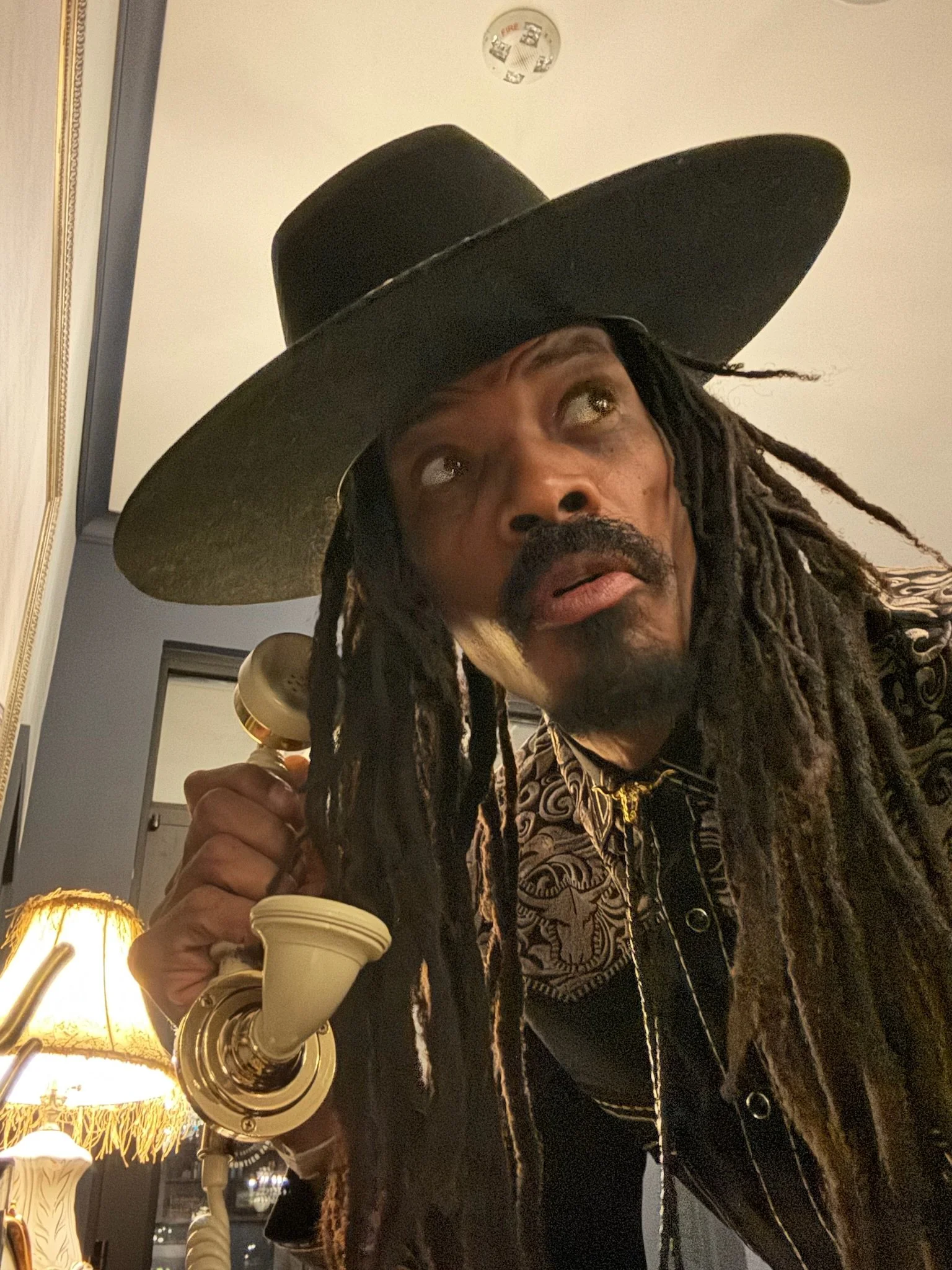 Man with long dreadlocks wearing a large black wide-brimmed hat, holding a vintage cream-colored telephone to his ear, standing in a room with a lamp and decorative trim.
