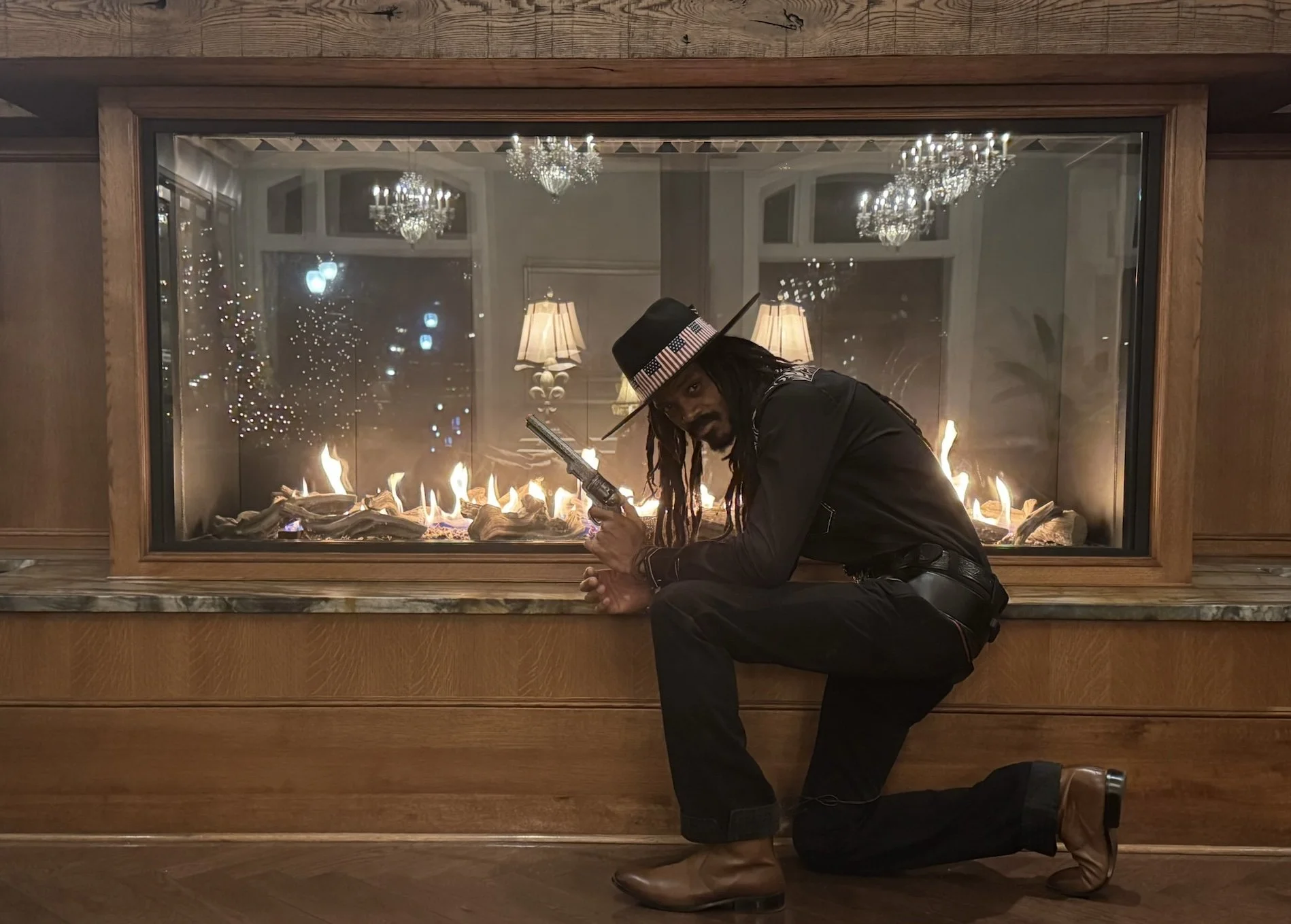 A man dressed in black leather outfit with a cowboy hat, sitting on one knee in front of a fireplace with flames, holding a revolver, in a room with chandeliers and lamps reflected in a glass window.