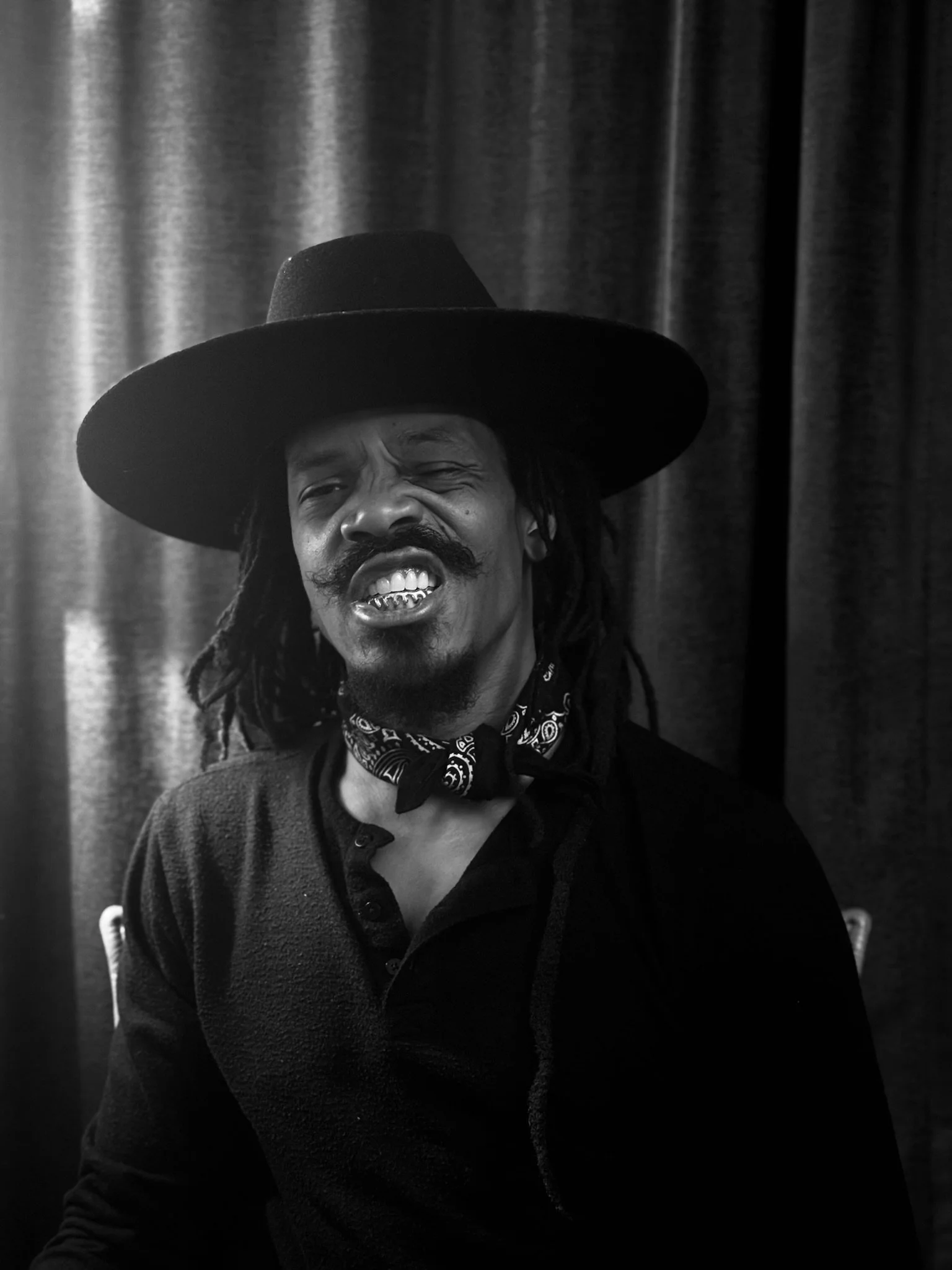 Black and white photo of a man with dreadlocks, wearing a large wide-brimmed hat, a bandana around his neck, and a dark shirt, standing against a curtain background.