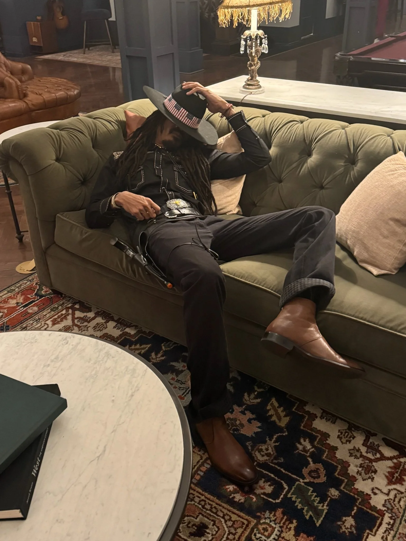 A man with dreadlocks is sitting on a green tufted couch, wearing a black jacket and jeans, holding a wide-brimmed hat with an American flag band. He is leaning back with one arm behind his head and his legs stretched out, one foot on the floor and t