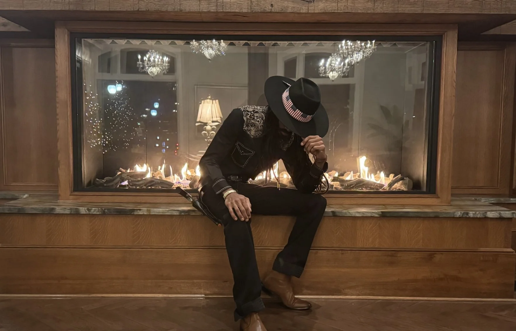 Person sitting on a wooden hearth in front of a glass fireplace, wearing a black hat with an American flag band, black outfit with embroidered shoulders, brown boots, and holding their face with one hand, indoor setting with chandeliers and lamps ref