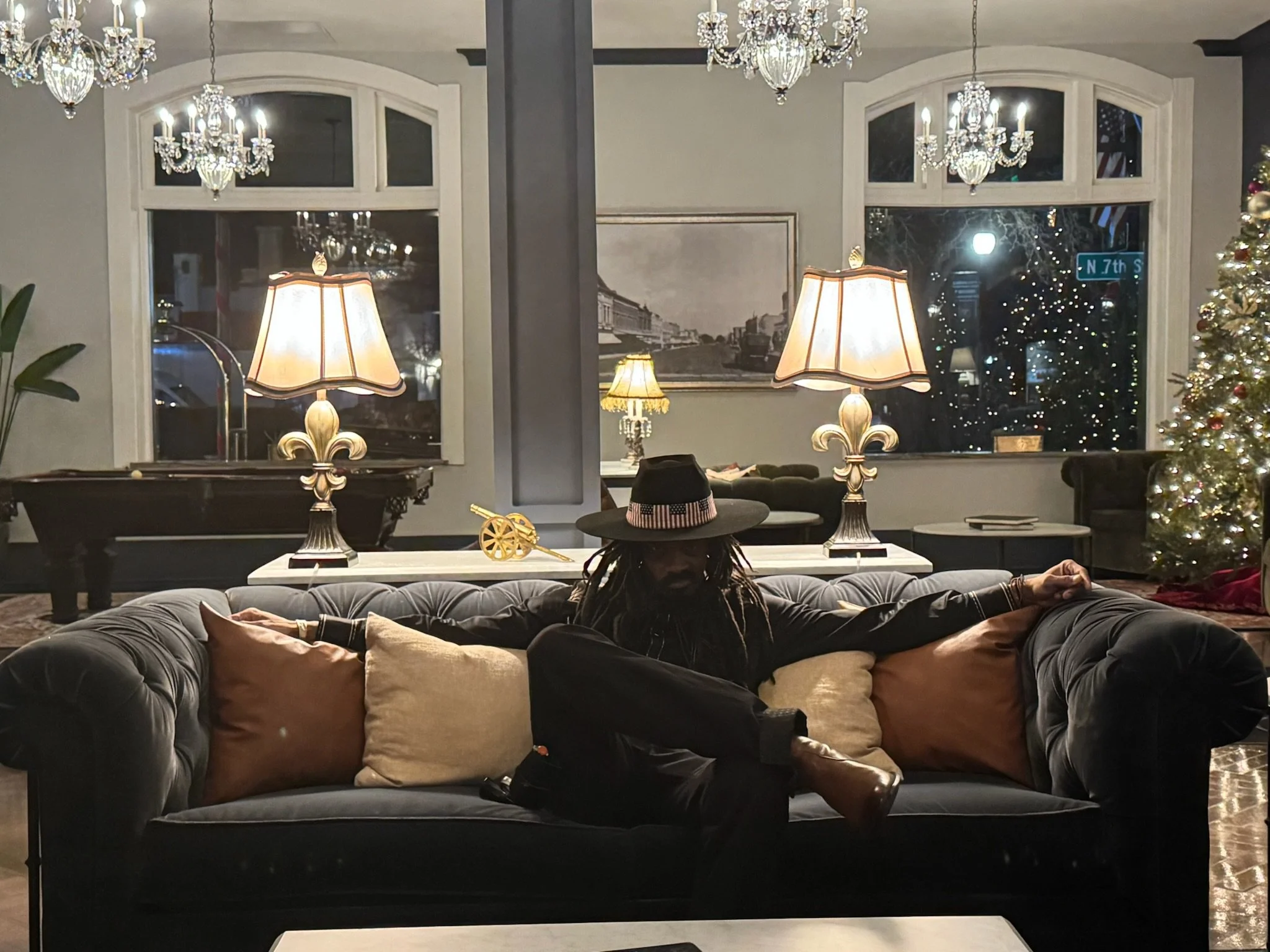A man with dreadlocks sitting on a dark gray velvet sofa with beige and light brown pillows, wearing a wide-brimmed hat and dark clothing, in a cozy living room decorated for Christmas with a lit Christmas tree on the right and lamps, chandeliers, an