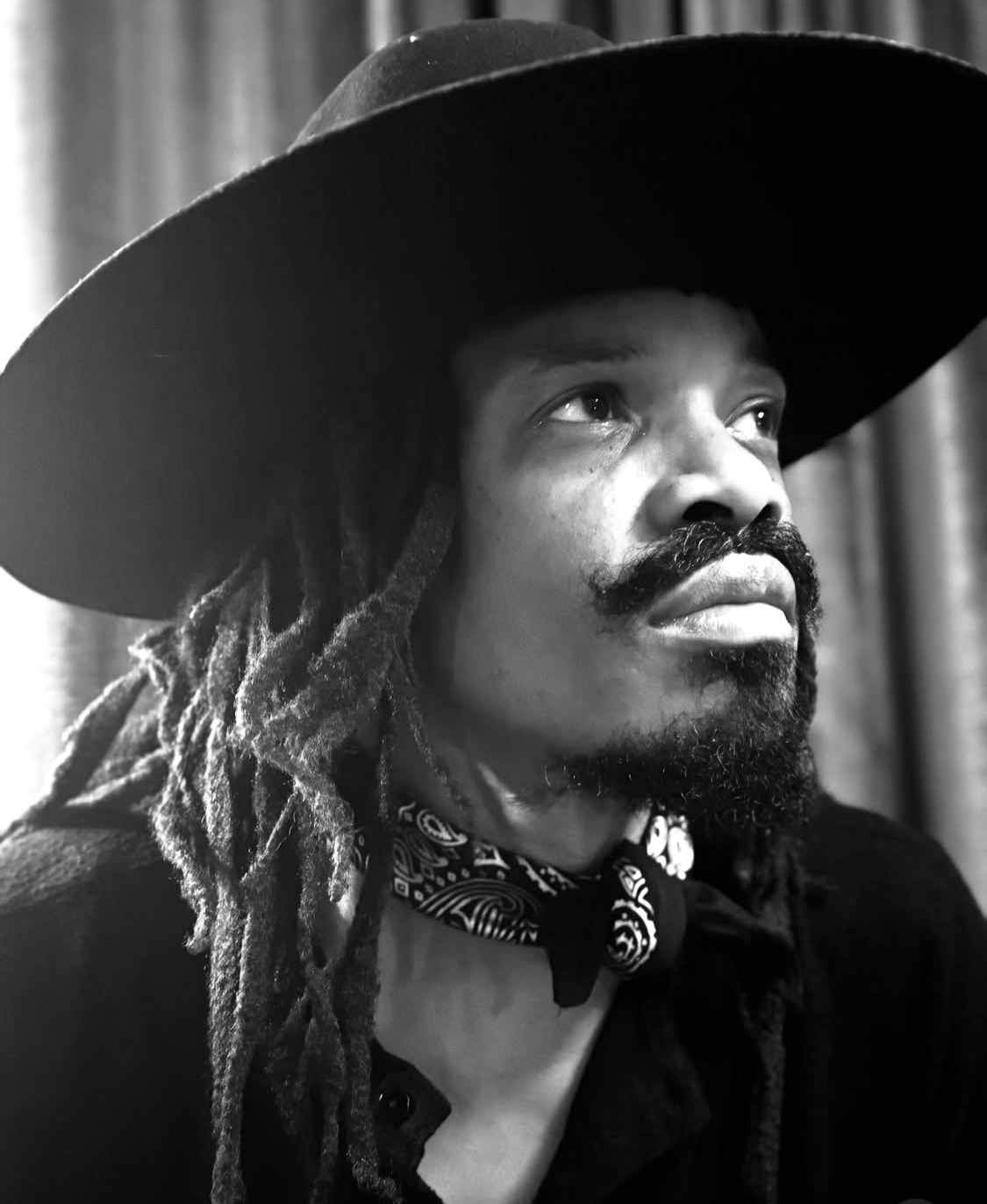 A man with dreadlocks and a mustache, wearing a wide-brimmed hat, a bandana around his neck, and a jacket, looking slightly upward in a black-and-white photo.
