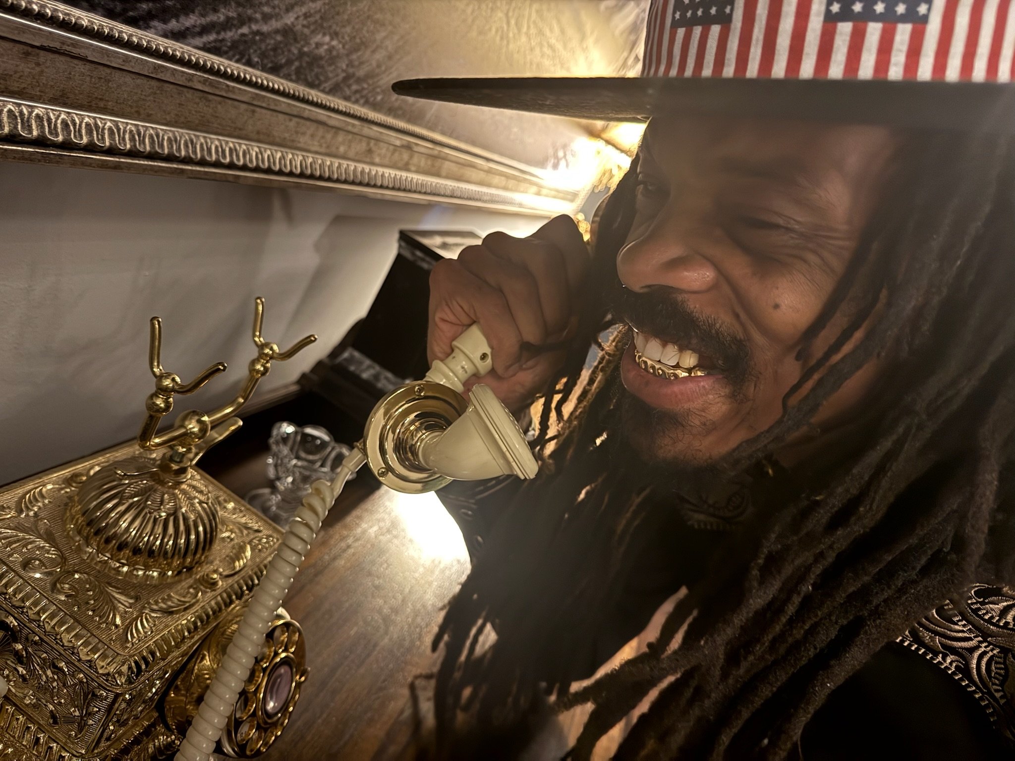 A man with dreadlocks, a gold grill, and a beard is smiling and talking on a vintage cream-colored rotary telephone on a wooden table. There is a decorative gold desk clock and a gold jewelry holder nearby. An American flag is in the background.