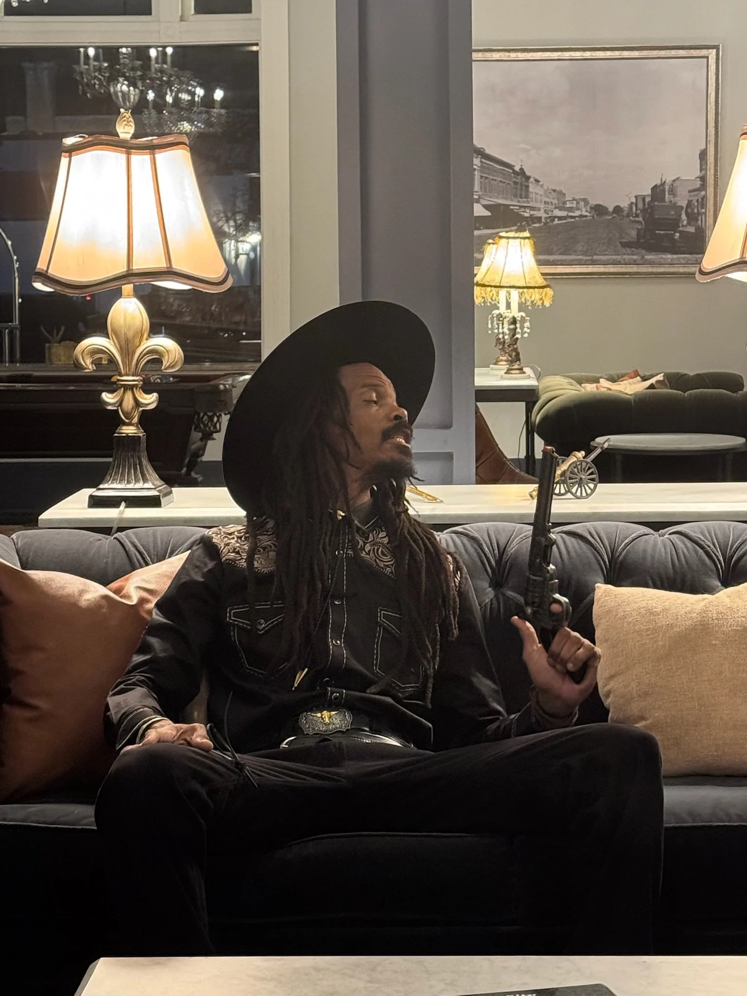A man with dreadlocks, wearing a large black hat, black shirt with ornate embroidery, and black pants, sitting on a dark gray sofa with beige and brown pillows in a living room, holding a black handgun.