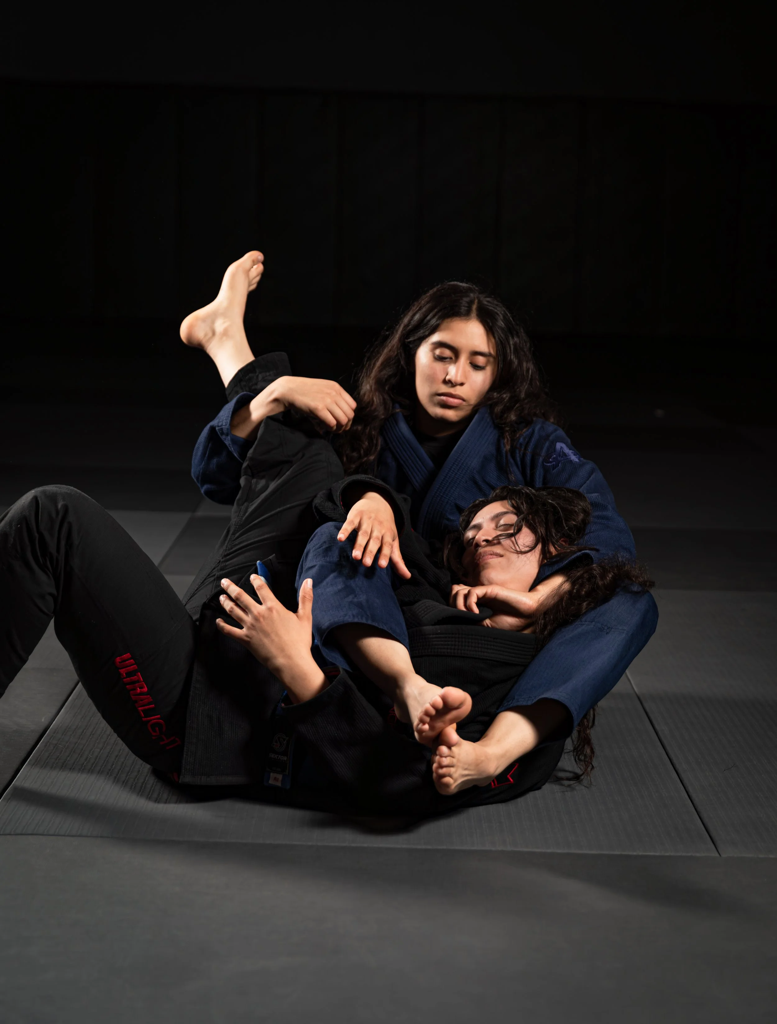 Two women practicing Brazilian Jiu-Jitsu on a black mat, one woman with long dark hair in a blue gi on top of a woman with curly hair in a black gi.